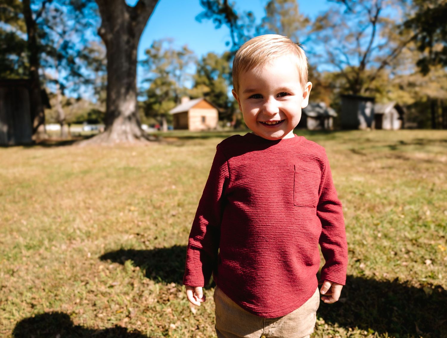 Young child in red sweater smiling outdoors on fall day.