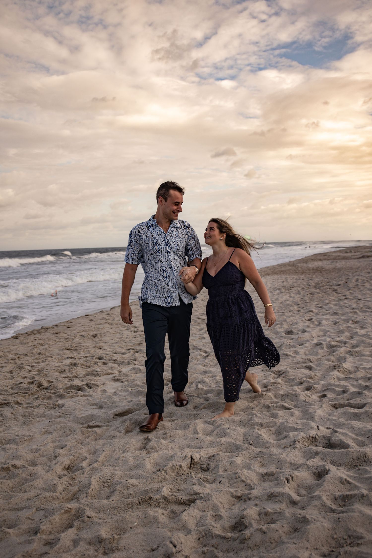 A couple walks together on a sandy beach at sunset with waves crashing in the background.