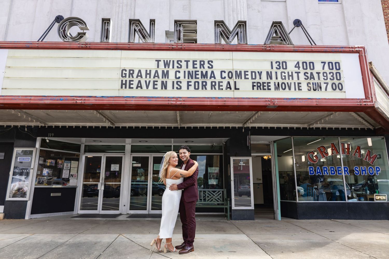A couple embraces in front of a vintage cinema marquee showing movie titles.