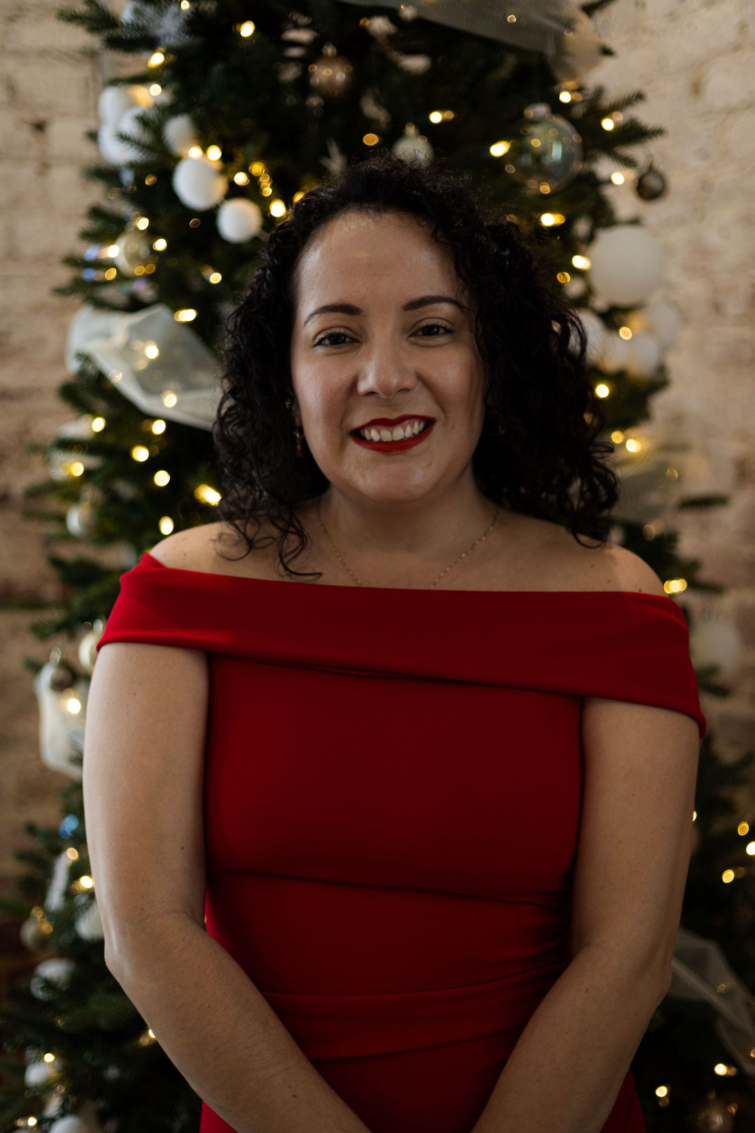 Woman in red off-shoulder dress smiles brightly against Christmas tree backdrop with twinkle lights.