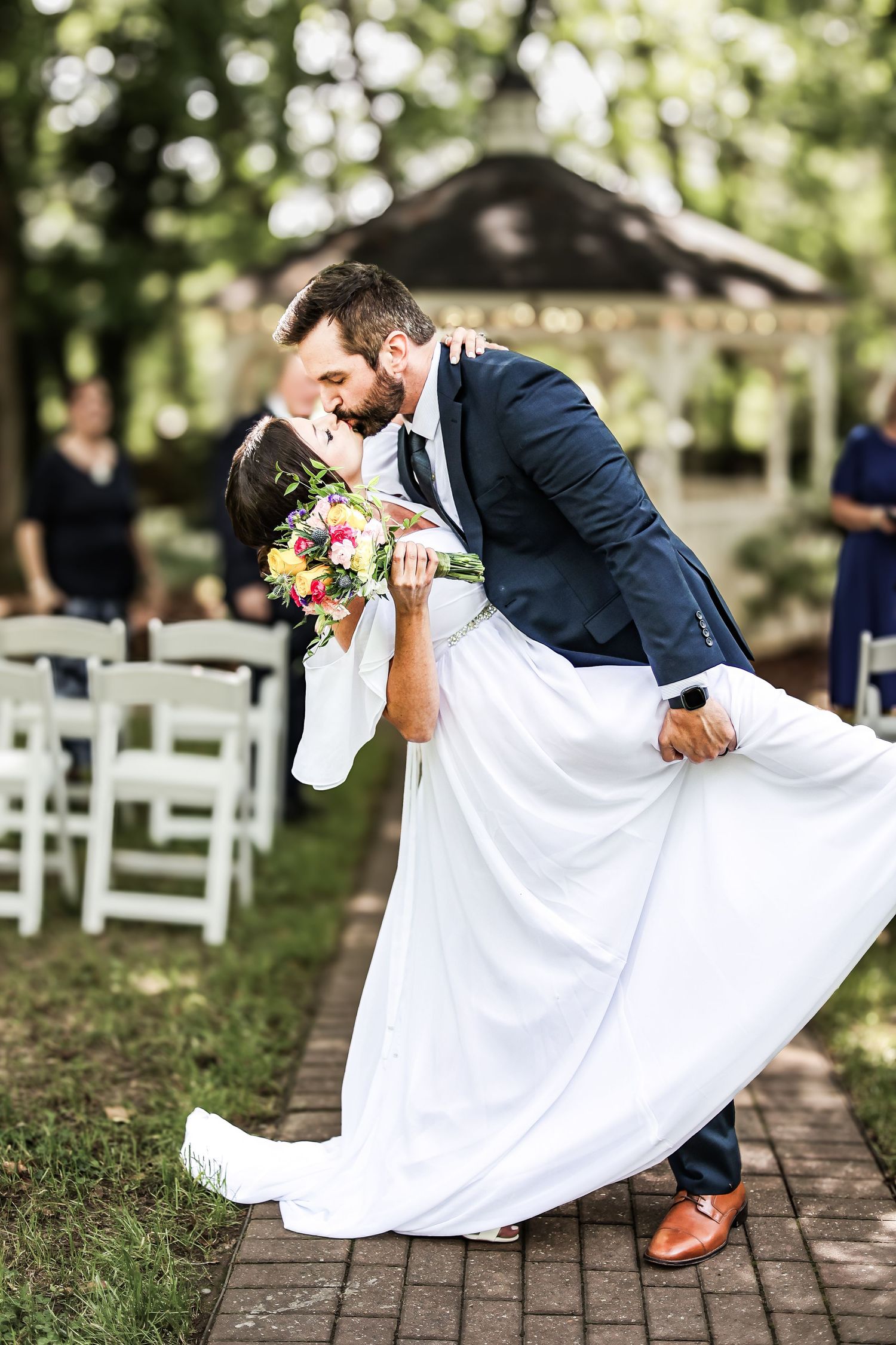 A wedding couple shares a romantic dip kiss on a brick path with white chairs and a gazebo in the background.