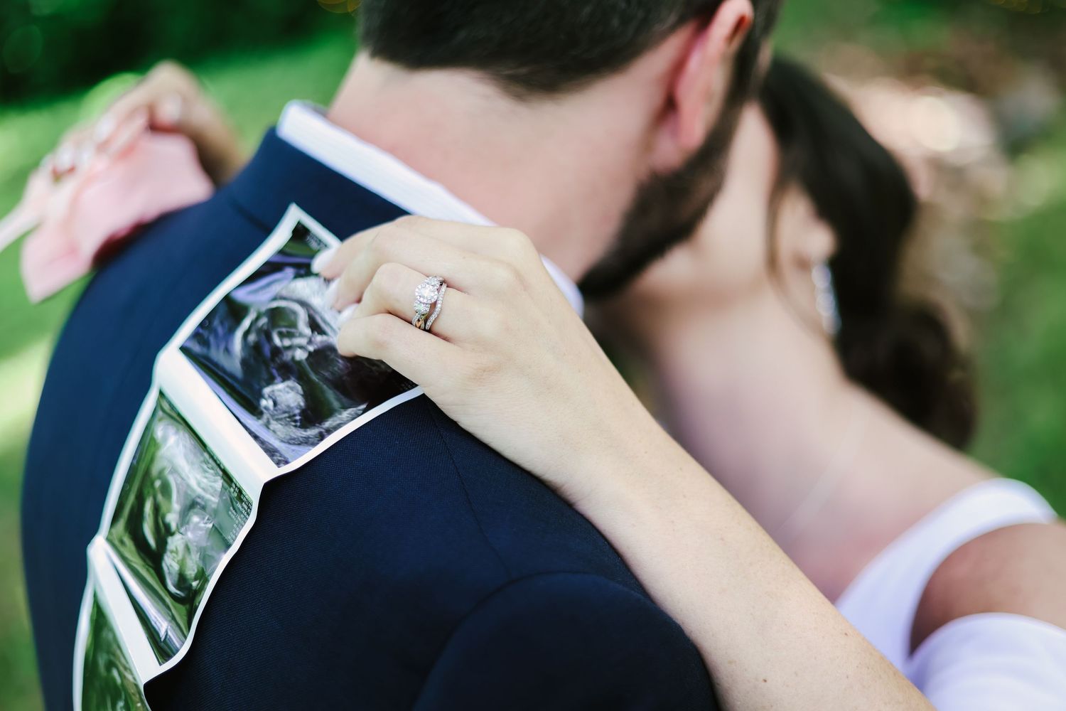 A couple embraces while holding ultrasound photos against a navy suit jacket outdoors.
