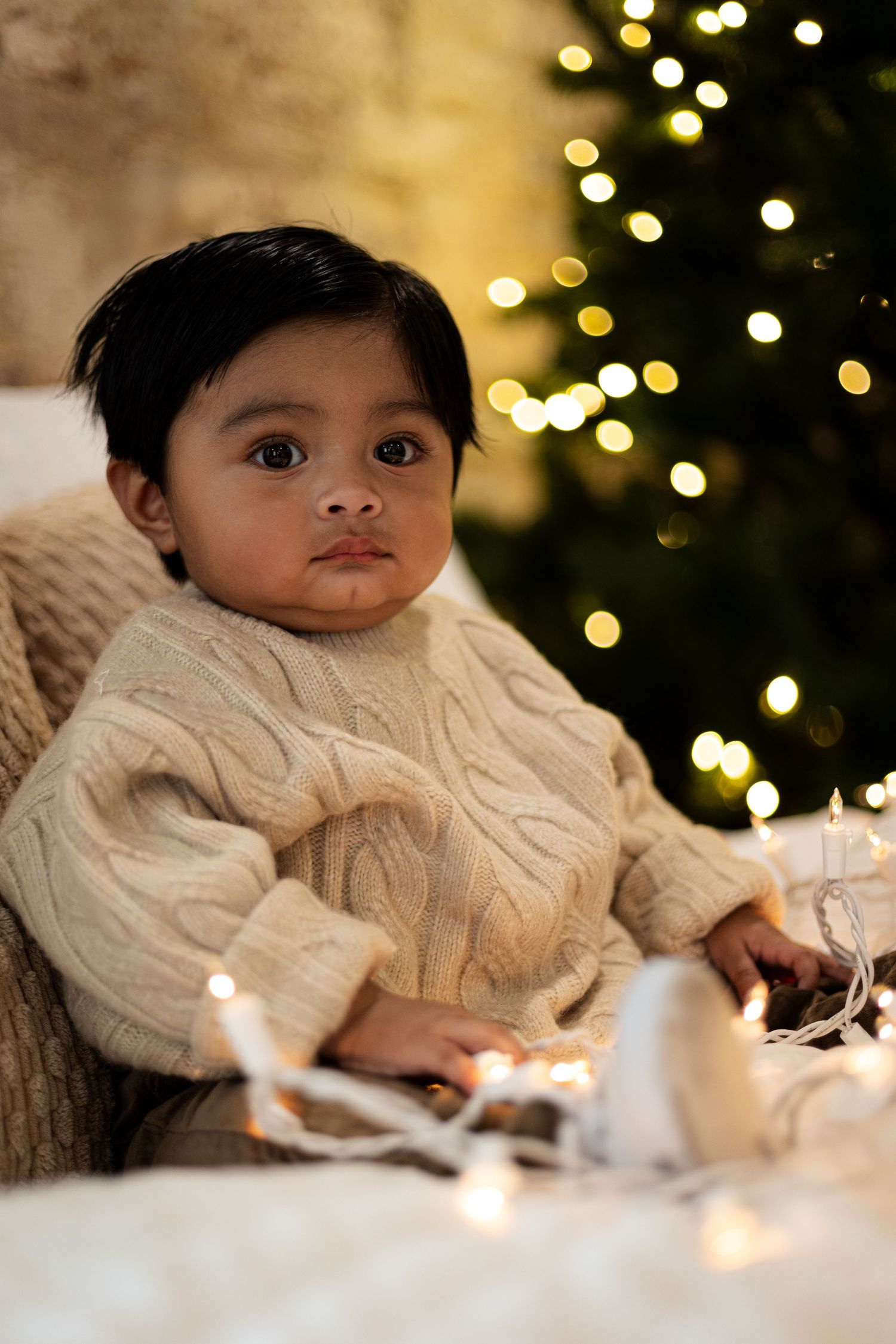Child in cream cable knit sweater sitting by Christmas tree with twinkling lights.