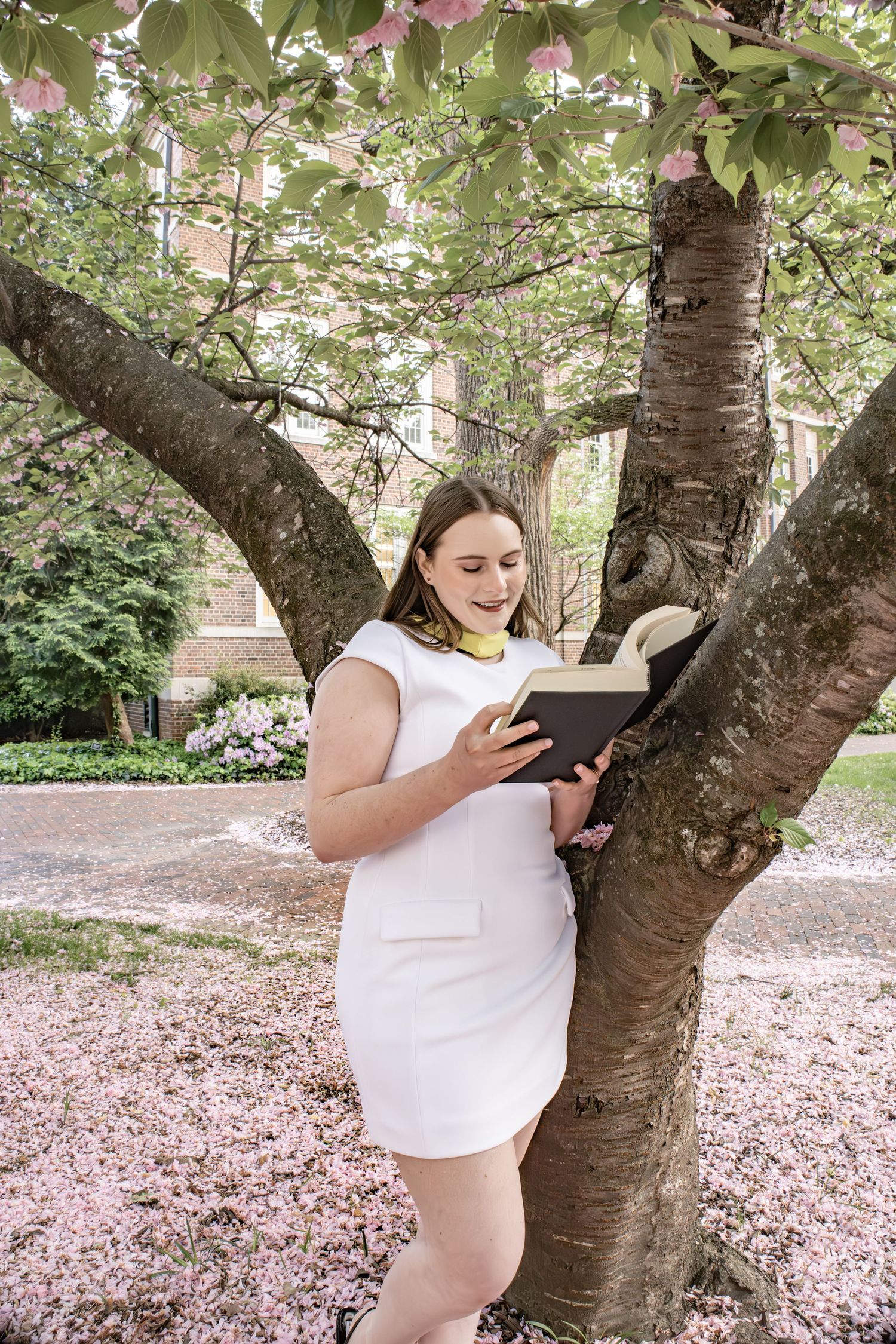 Person reading book while standing against cherry blossom tree.