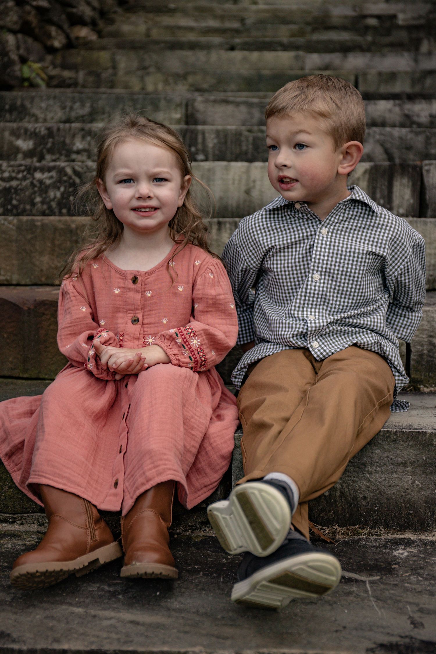Two young children sit together on stone steps wearing coordinating pink and gingham outfits in vintage style.