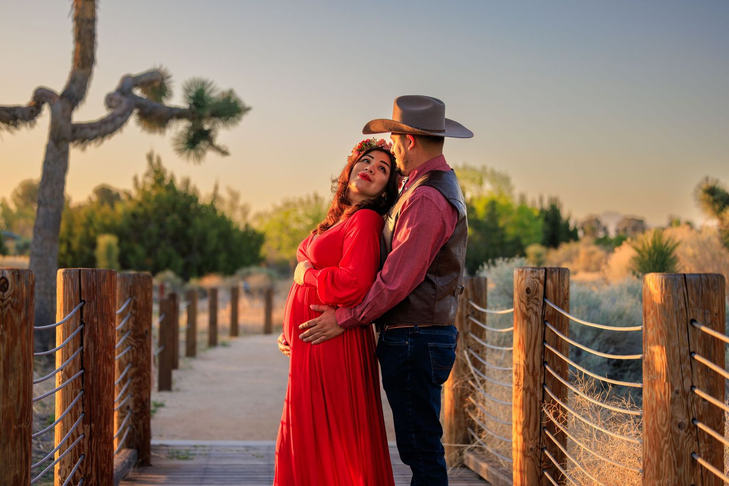 Romantic couple in western attire embracing on wooden bridge at sunset.