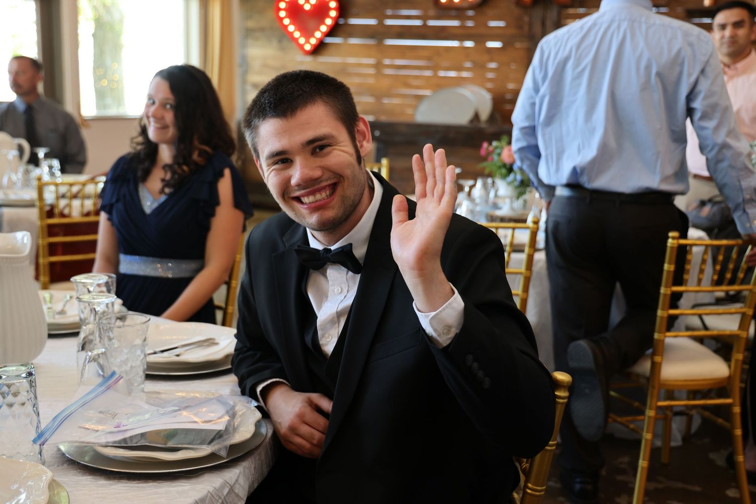 A guest in a black tuxedo waves while seated at an elegantly set wedding reception table.