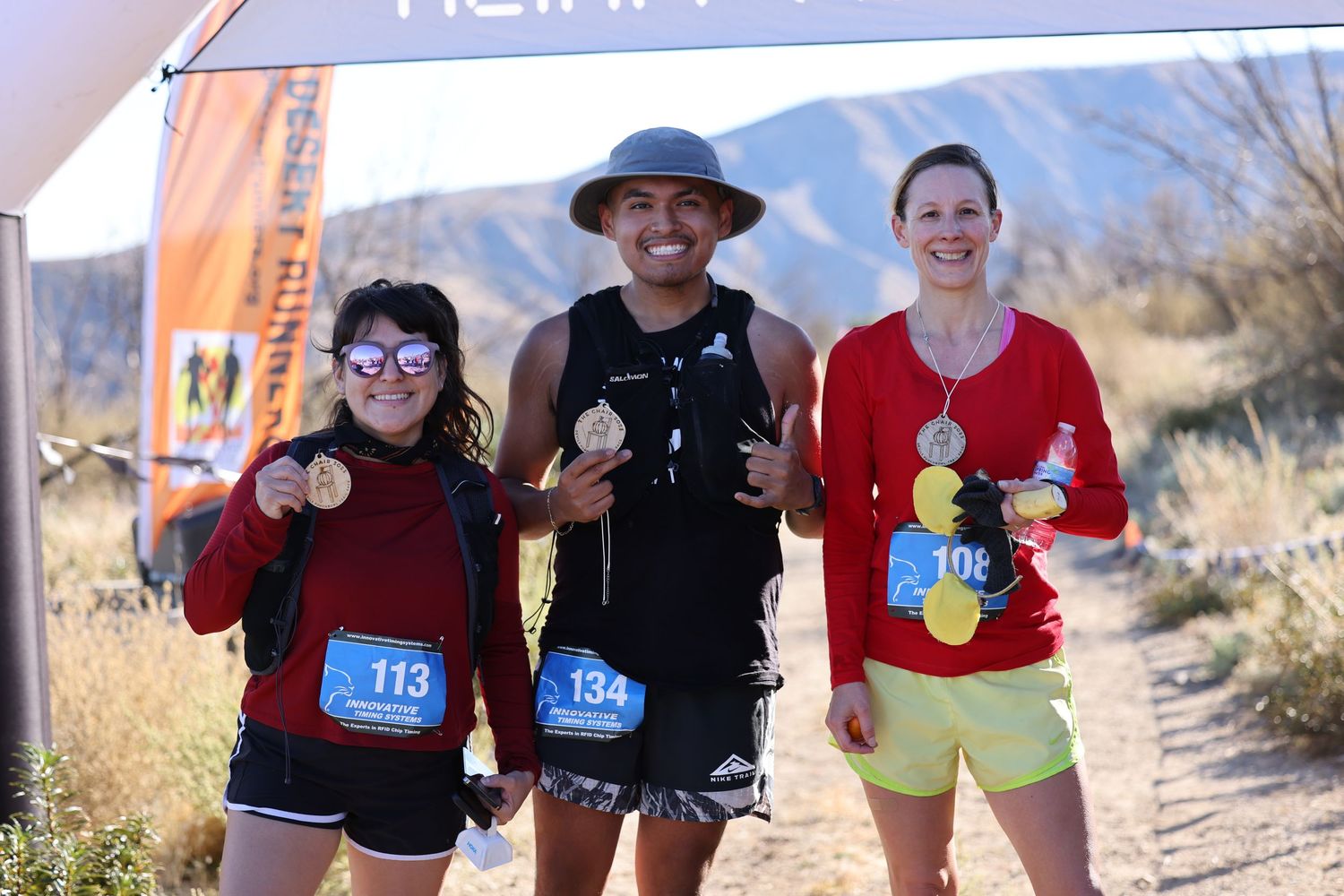 Three runners smiling and posing together under a finish line banner at a trail race.