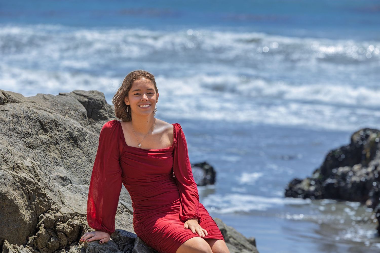 A person in a red dress sits on coastal rocks with ocean waves crashing in the background.
