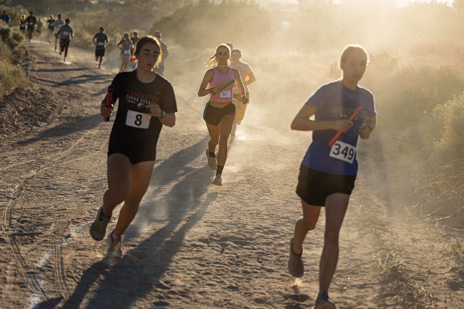 Runners compete on a dusty trail during golden hour as sunlight streams through the morning mist.