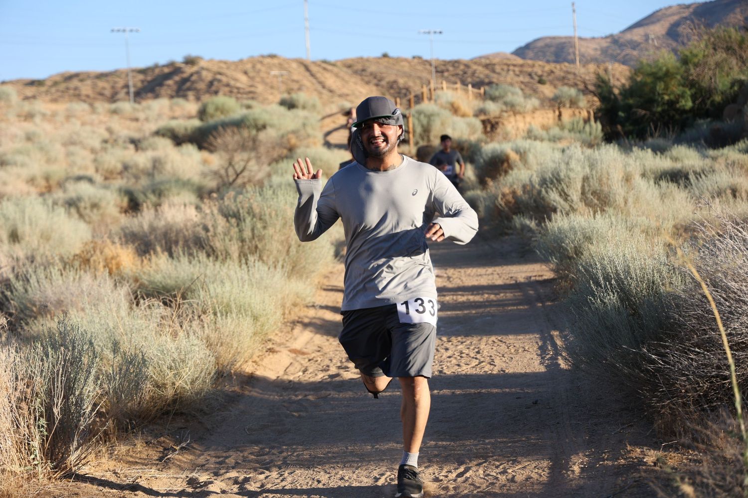 An enthusiastic runner waves while jogging on a desert trail with mountains in the background.