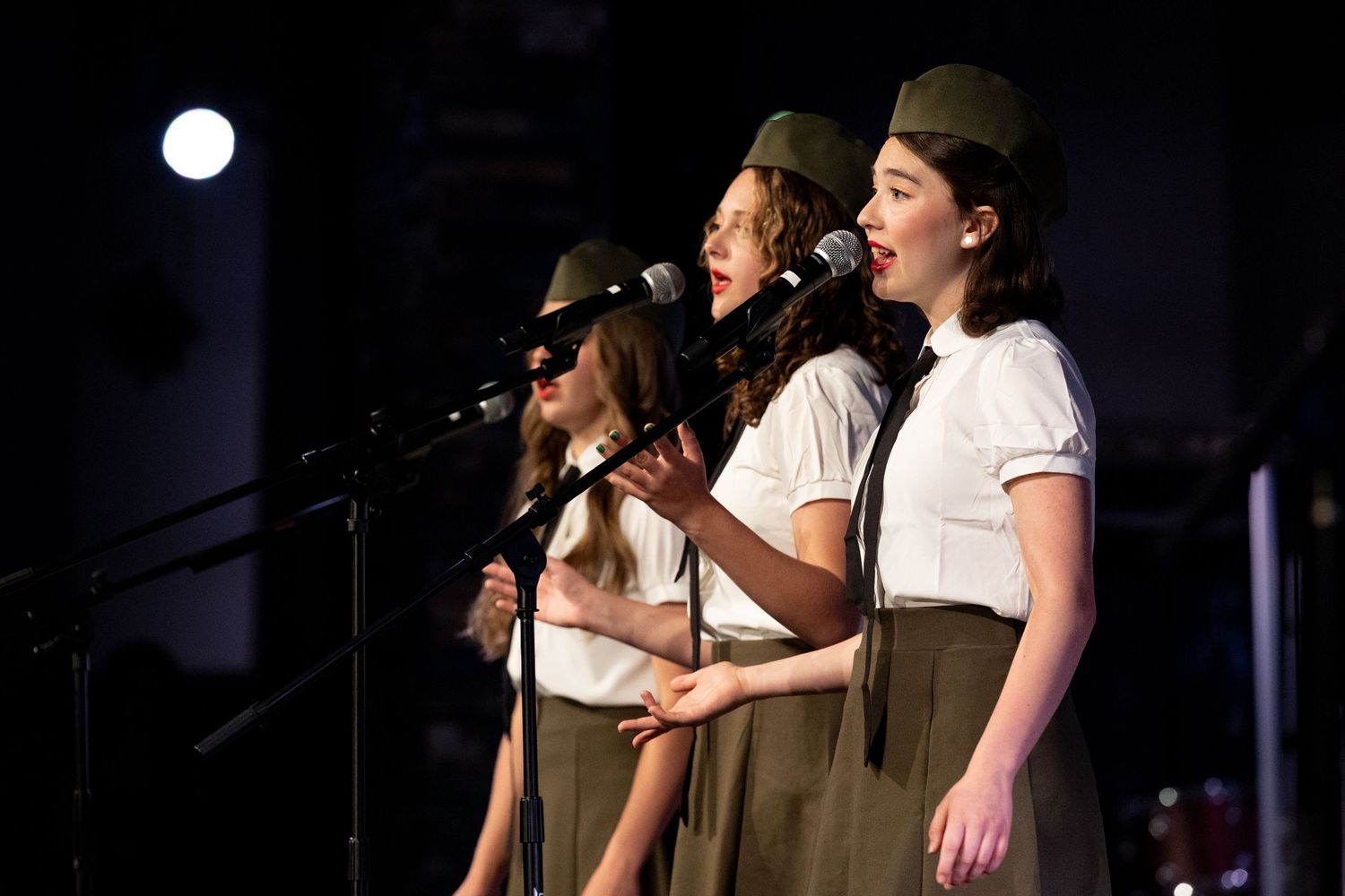 Performers singing in harmony at a microphone, dressed in vintage-style brown and white outfits on a dark stage.