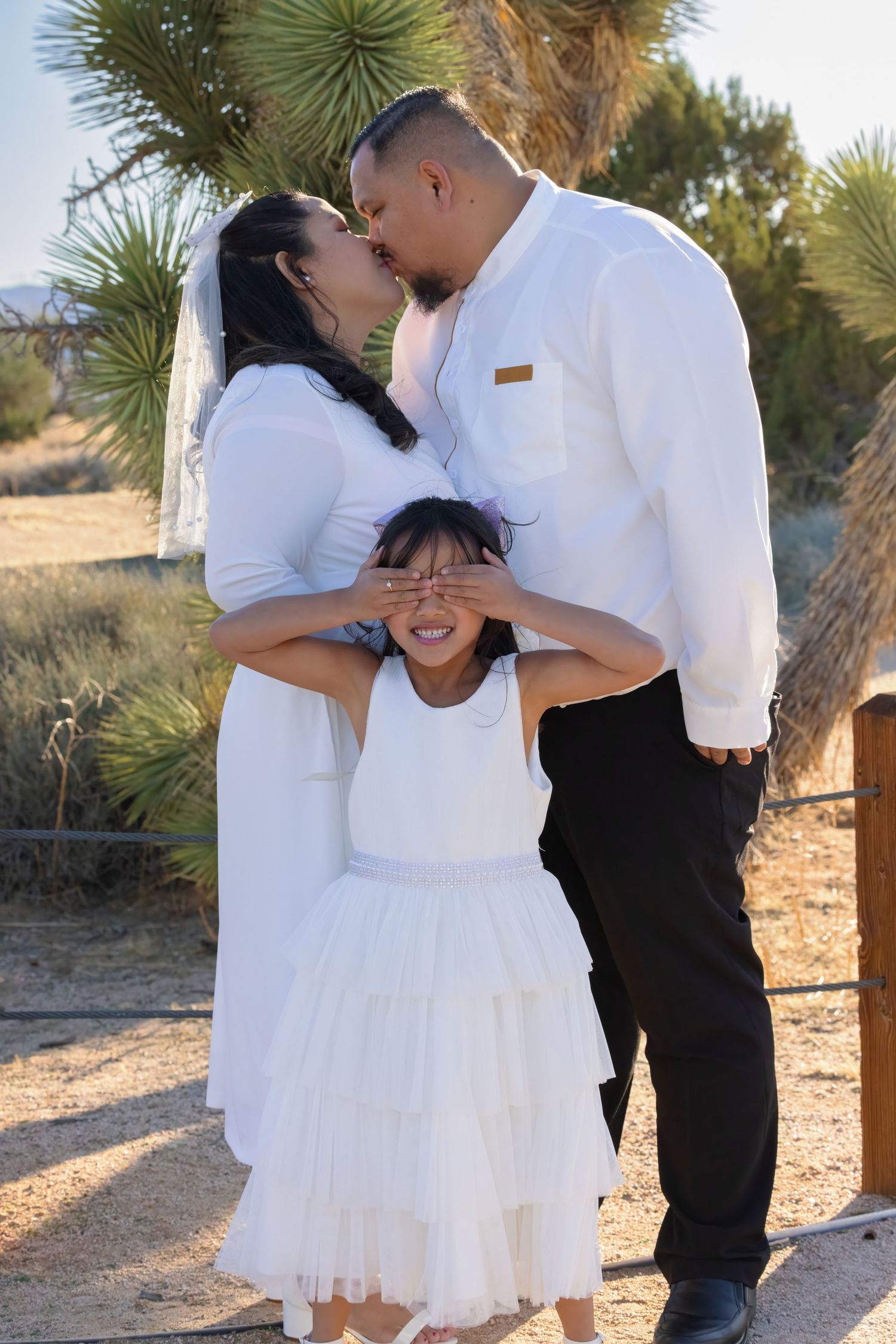 A sweet family photoshoot outdoors with parents kissing while child in white dress playfully covers their eyes.