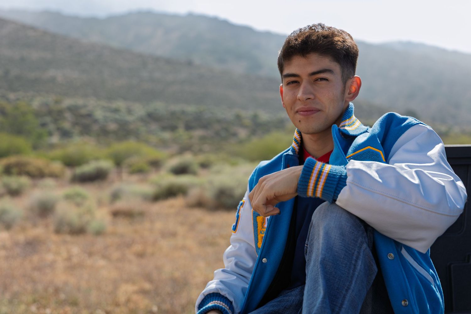 Young person in a blue and white jacket sits outdoors against a desert mountain landscape backdrop.
