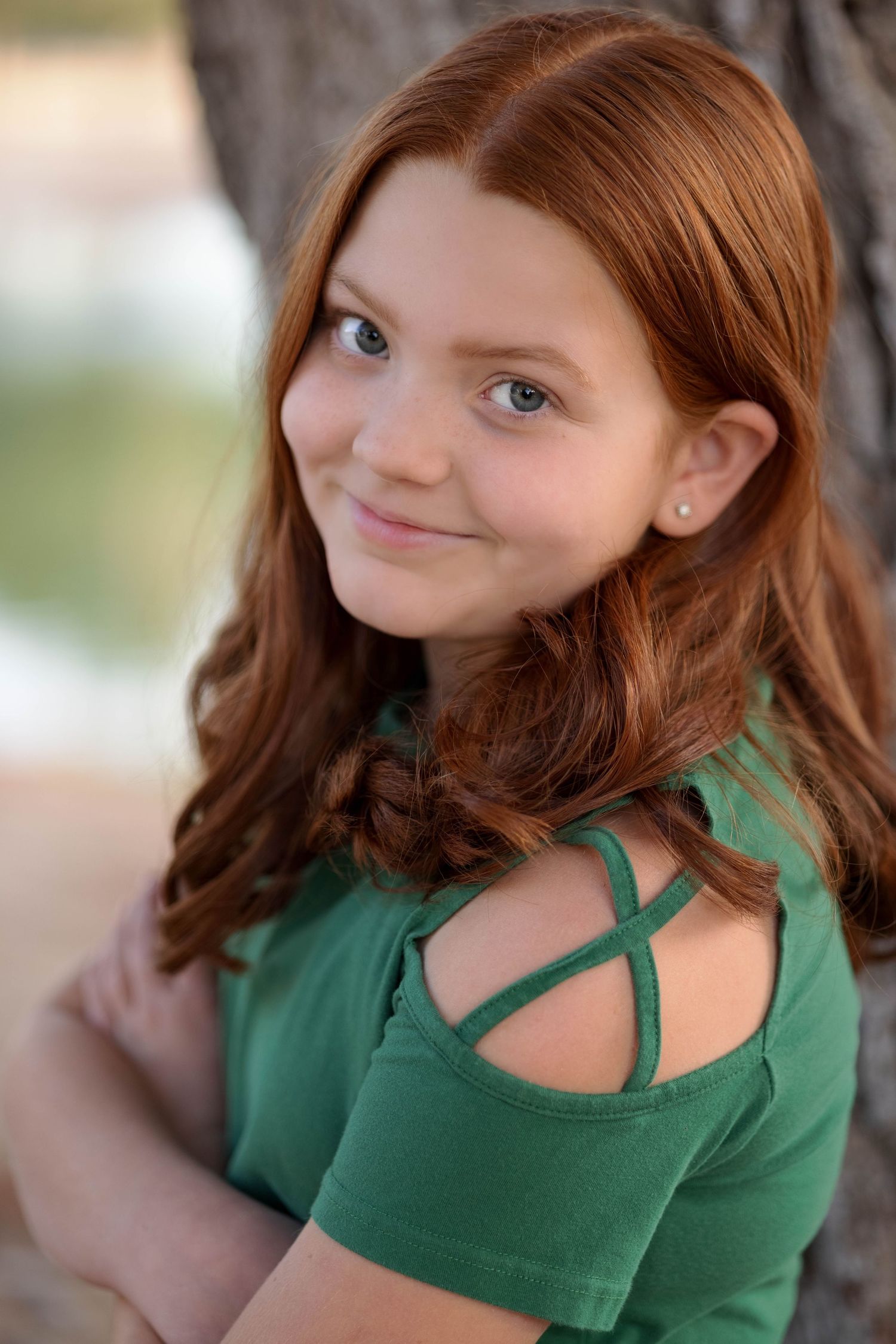 Young person wearing green top with criss-cross shoulder detail smiles warmly at camera in outdoor portrait.