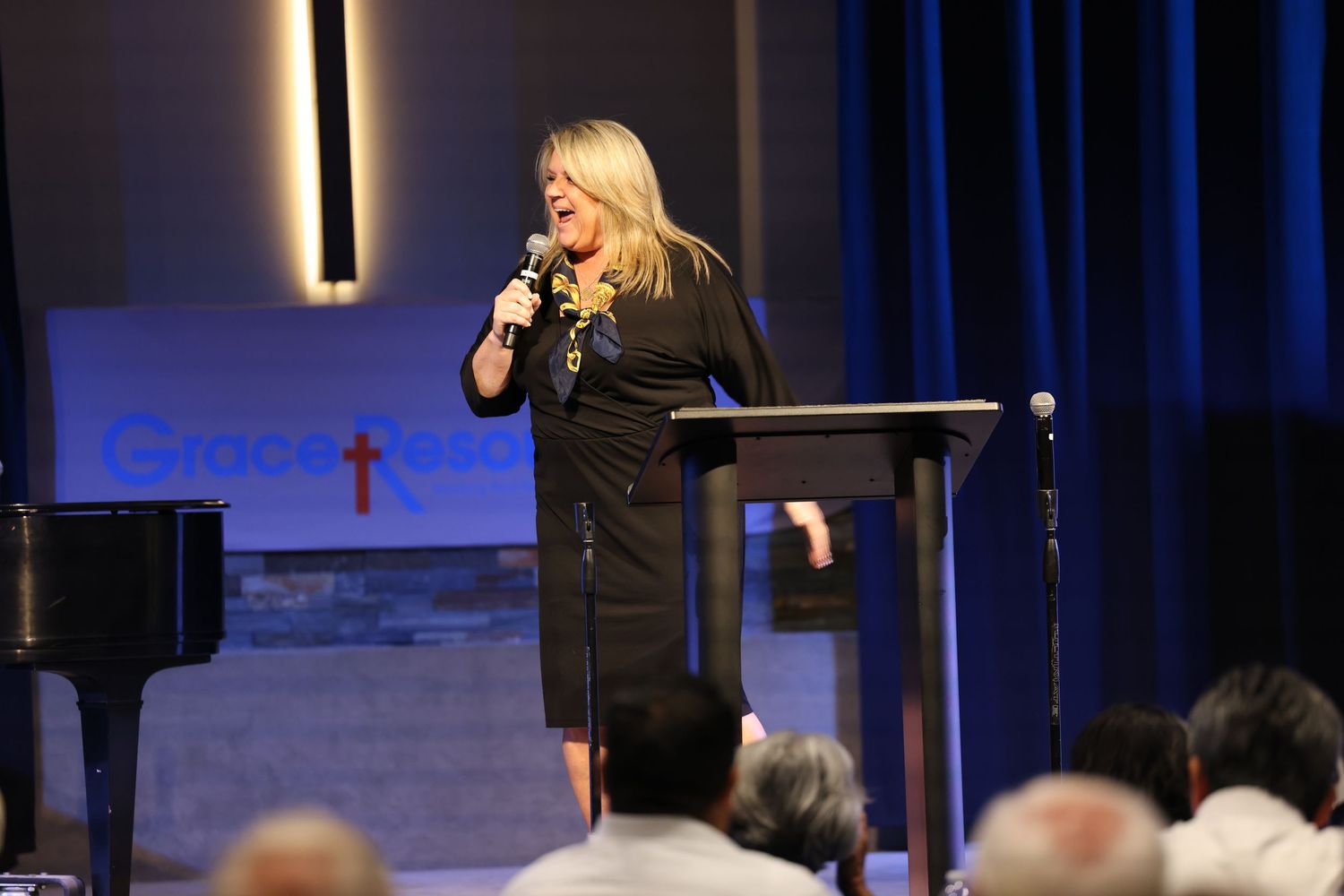 A performer speaking at a podium with a piano and illuminated cross visible on stage.