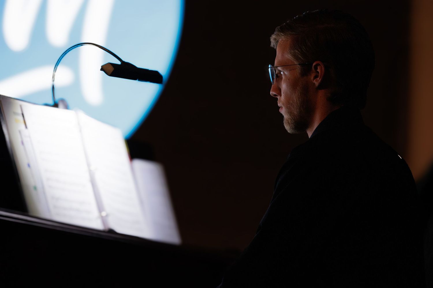 A person sits at a dimly lit desk reading sheet music illuminated by a small light.