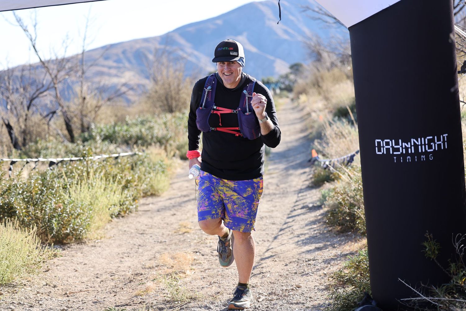 Trail runner in athletic gear jogs along a desert path with mountains in the background during a daytime run.