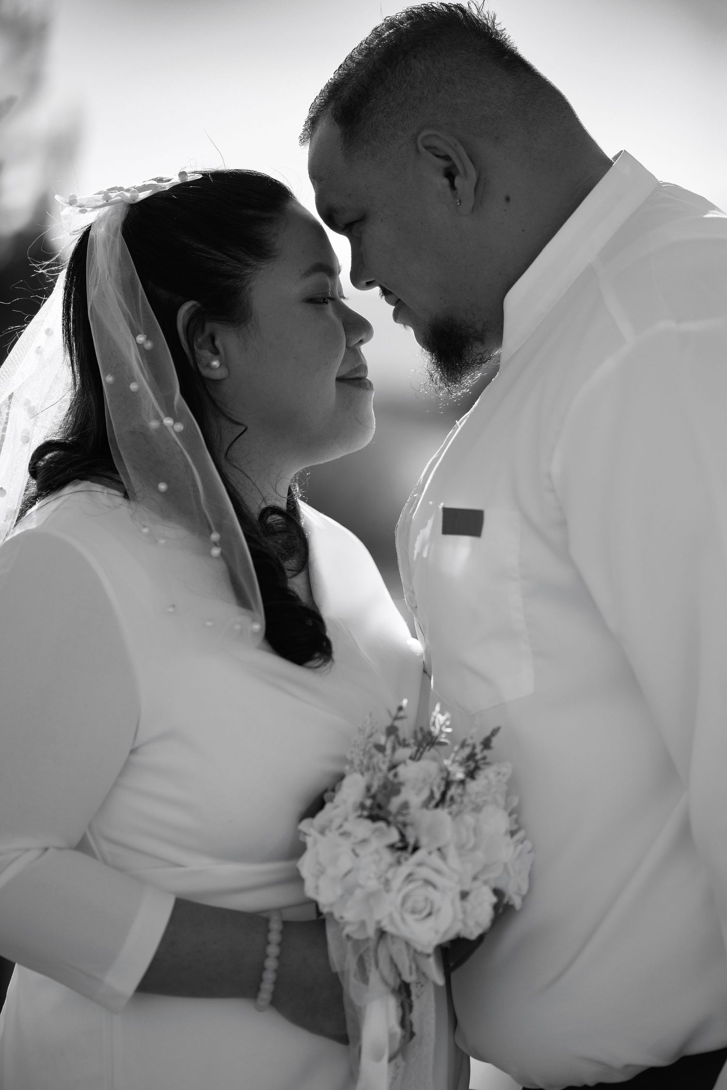 Intimate black and white wedding portrait showing tender moment between newlyweds with floral bouquet.