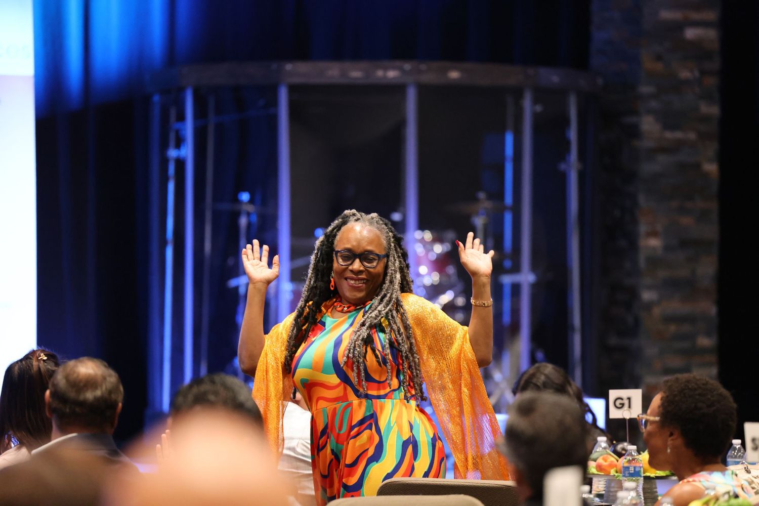 A person in colorful attire raises their hands while speaking at a conference.