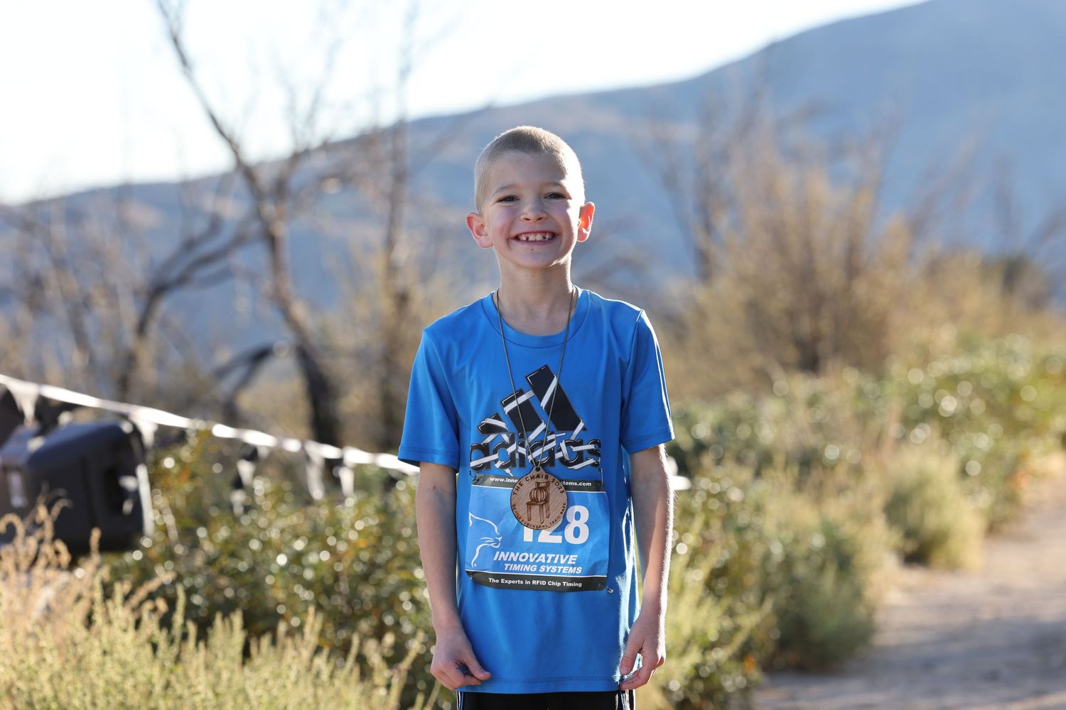 Child in bright blue t-shirt smiles while standing on a mountain trail.