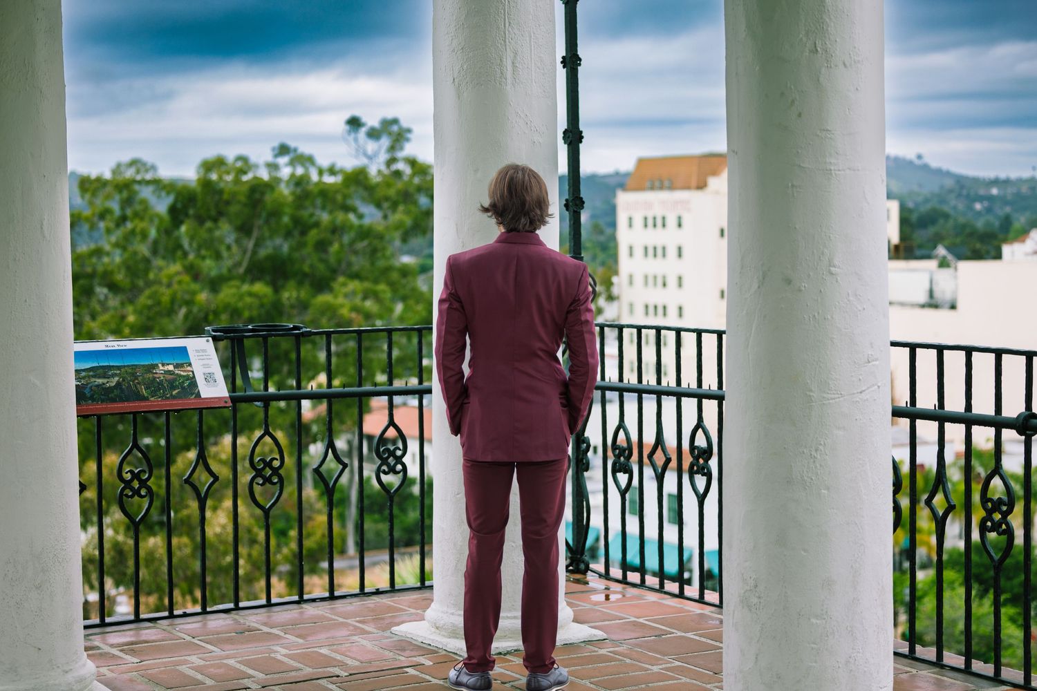 View through white columns of a balcony overlooking a tropical landscape with buildings and palm trees in the distance.