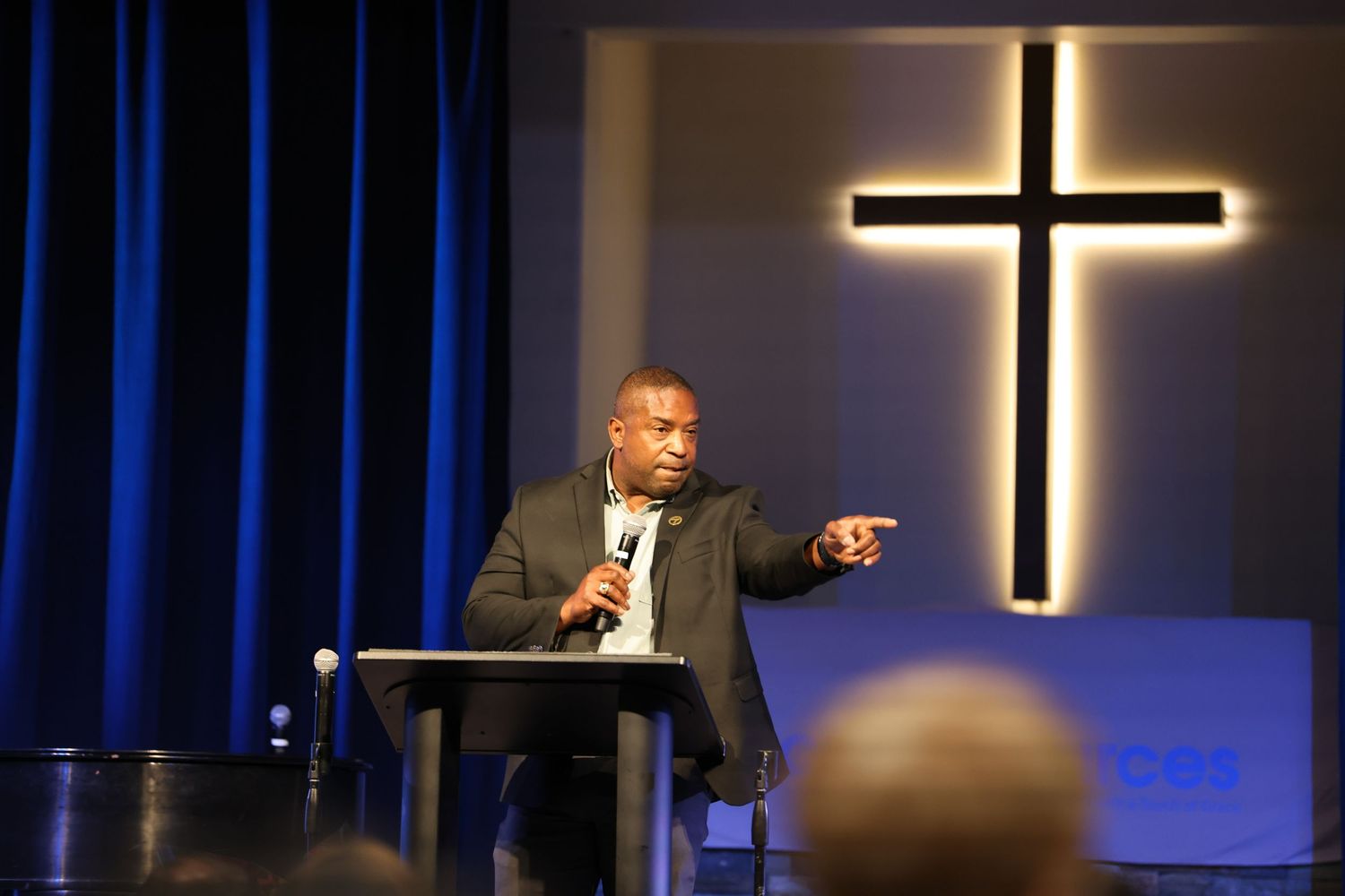 Speaker gesturing from church podium with illuminated cross on stage backdrop.