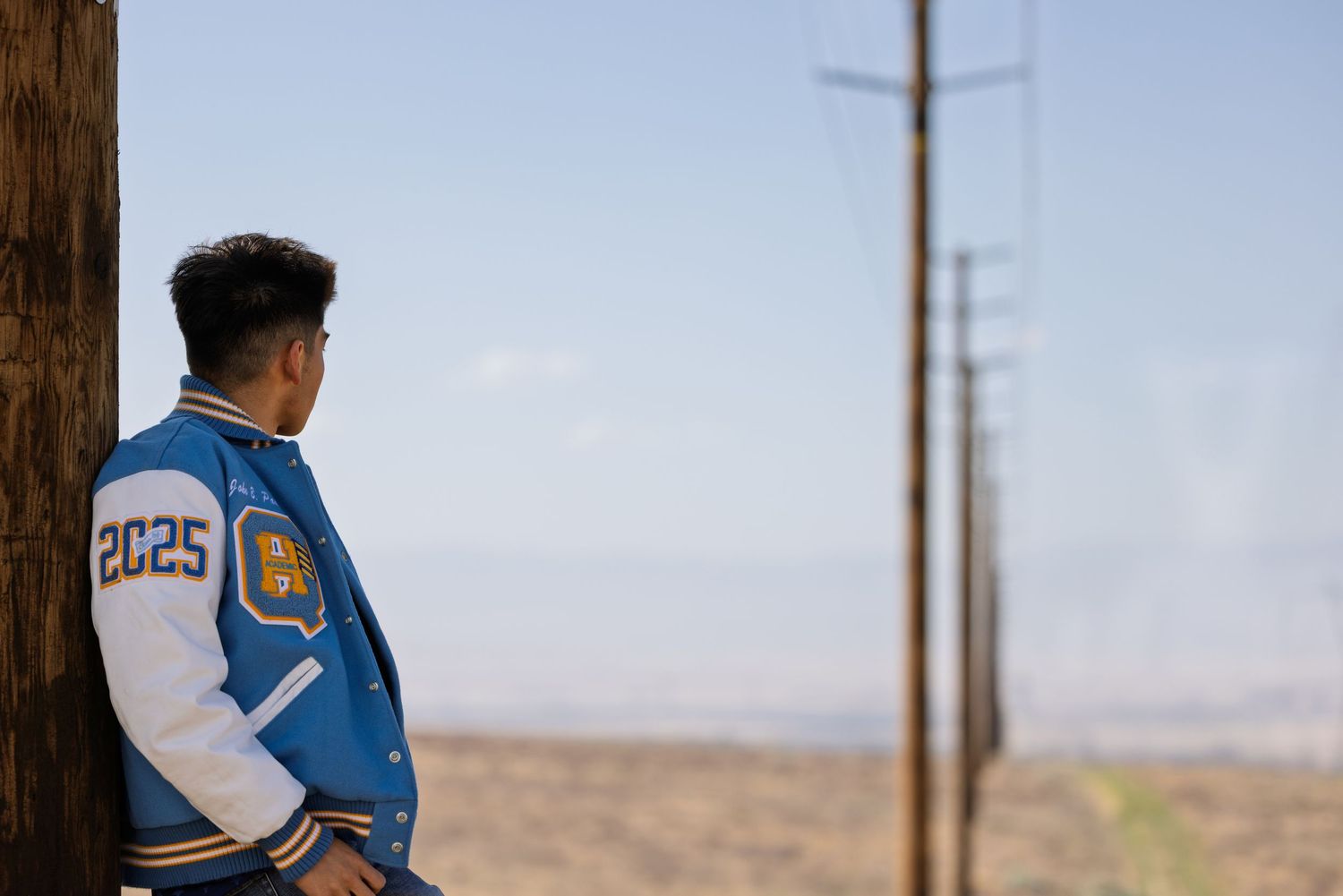 A person in a blue and white varsity jacket stands near a wooden pole overlooking a desert landscape.