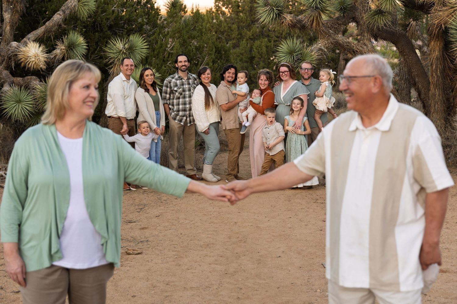 A group gathers outdoors at sunset to celebrate as two people reach out to hold hands on a dirt path.