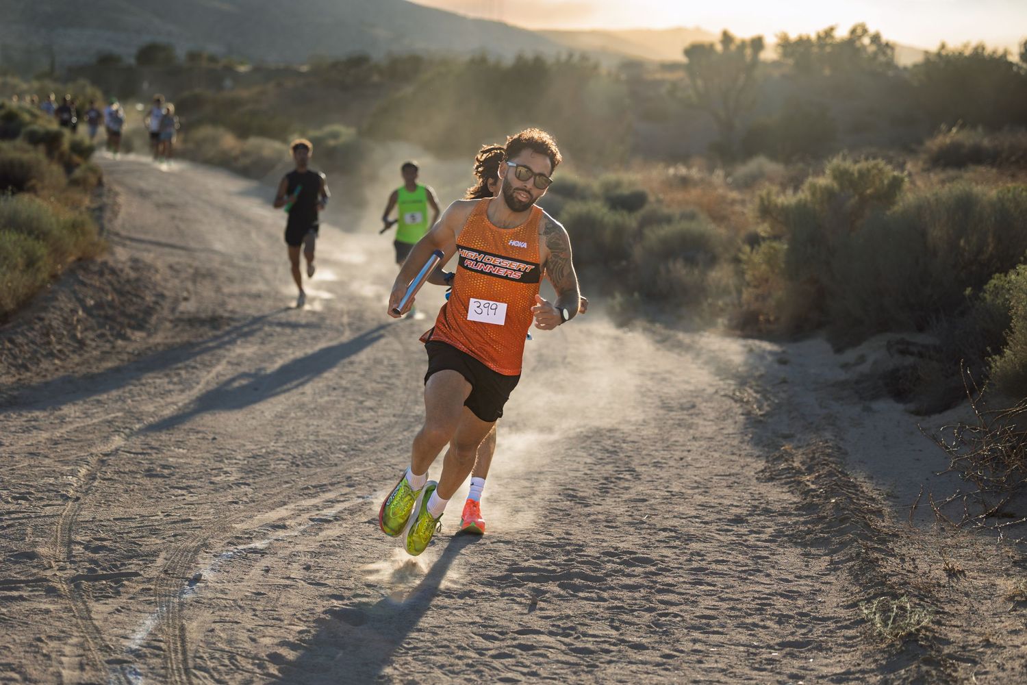 Trail runners racing on a dusty mountain path at sunset with golden light illuminating the rugged desert terrain.