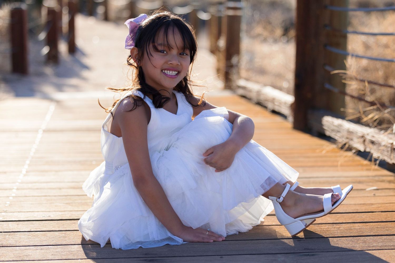 A child in a white dress and shoes sits playfully on a wooden deck.