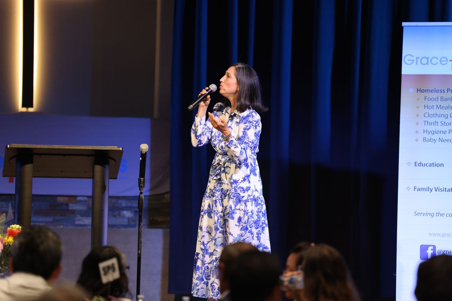 Woman in floral dress singing into microphone on church stage with cross display.