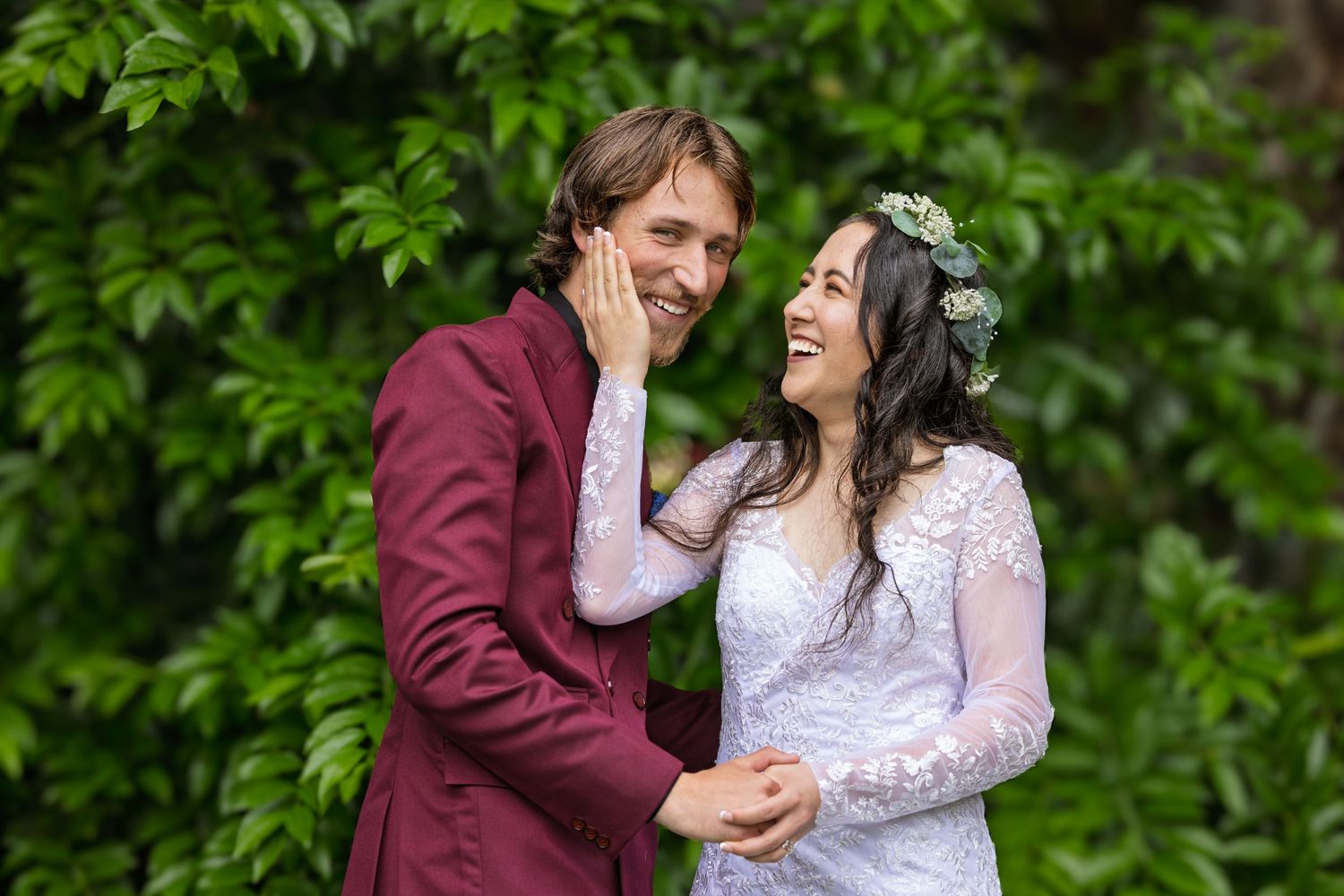 A couple shares a joyful moment while dressed in wedding attire with a burgundy suit and white lace gown against greenery.