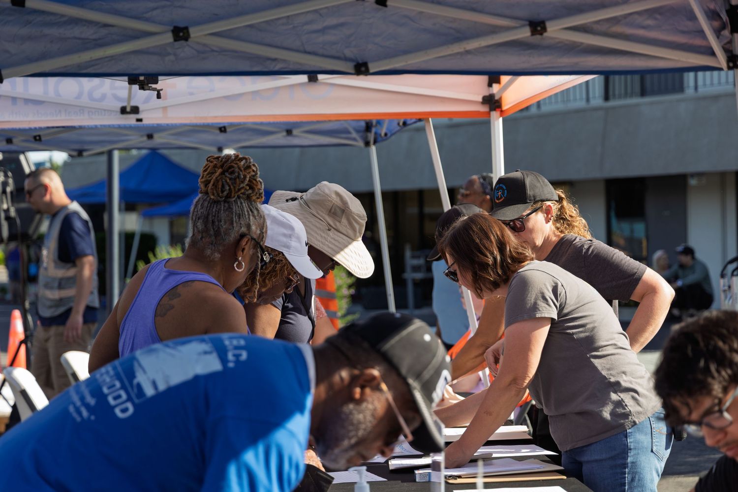 People gather under a white canopy tent at an outdoor event during a sunny day.