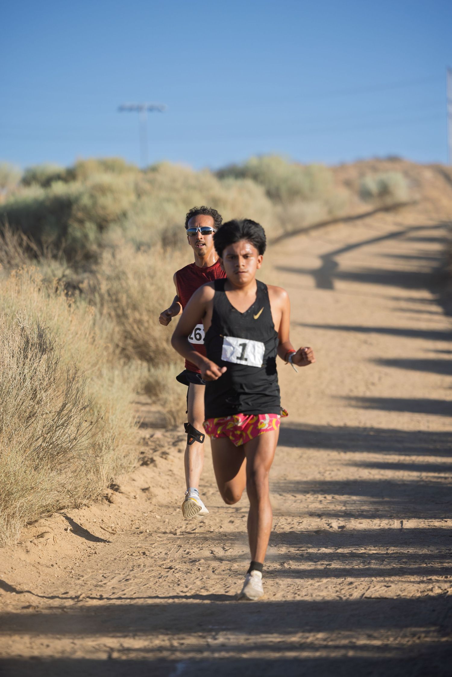 Trail runners competing on a dusty dirt path through dry grassland on a sunny day.