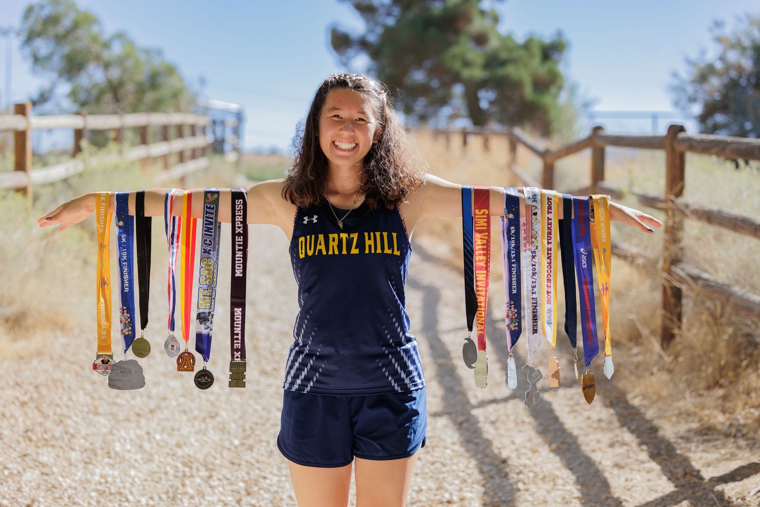 An athlete in a navy uniform displays multiple medals while standing on a dirt trail.
