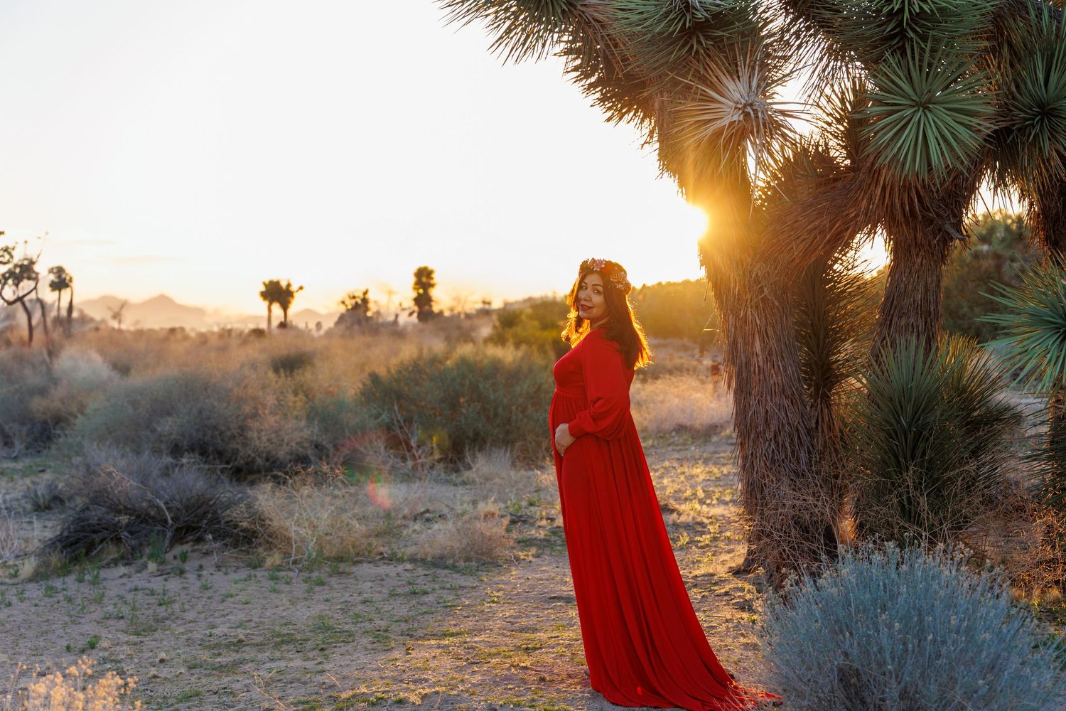 Woman in flowing red dress poses dramatically under Joshua tree at golden hour.