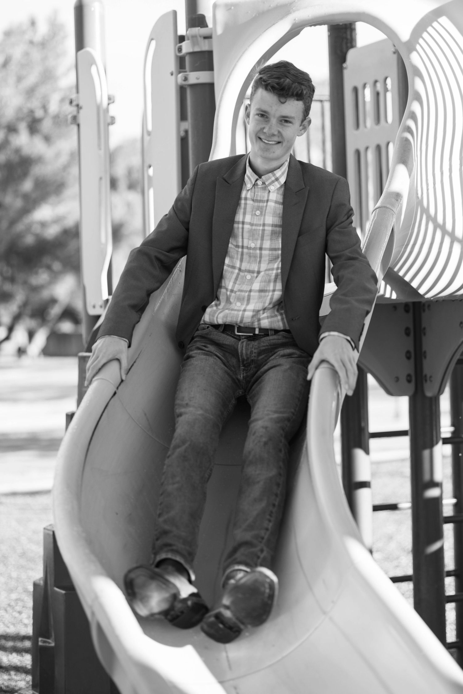 A playful black and white portrait of someone sitting on a playground slide wearing business casual attire.