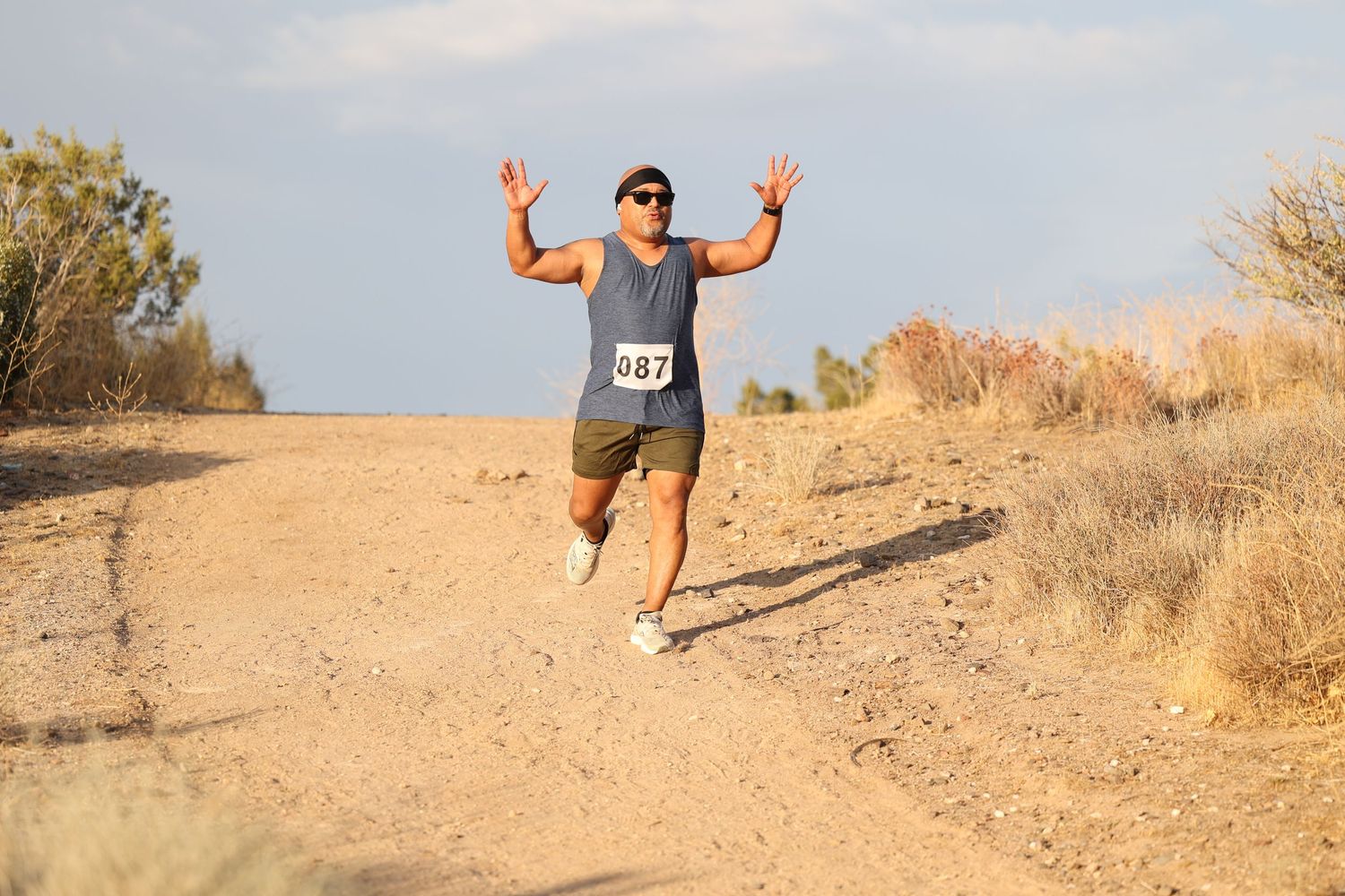 A runner gestures victoriously while running on a dusty desert trail.