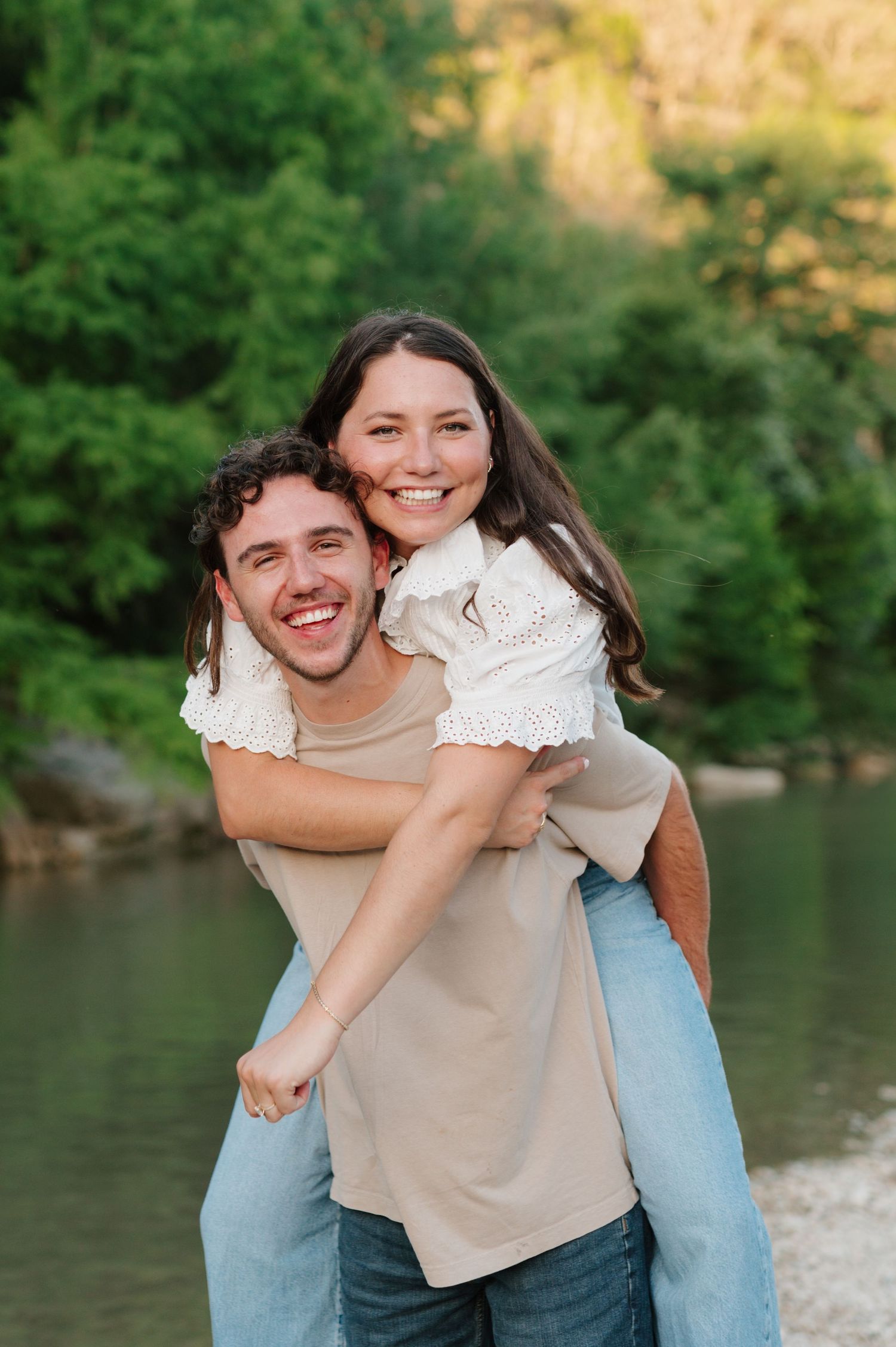 A playful couple shares piggyback rides and laughter by a scenic lake with greenery in the background.