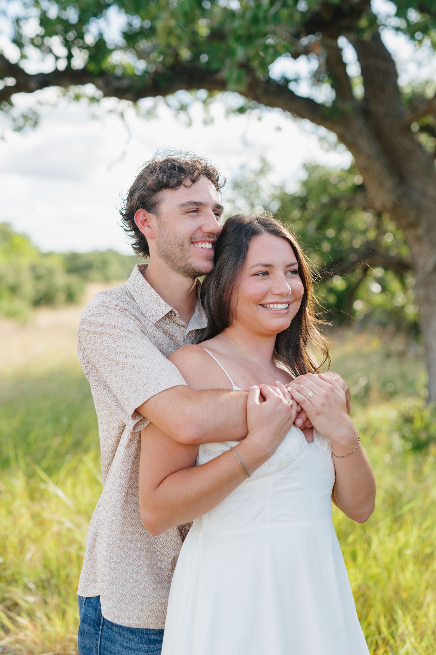 A romantic outdoor photoshoot in a sunny field with a couple embracing under a tree.