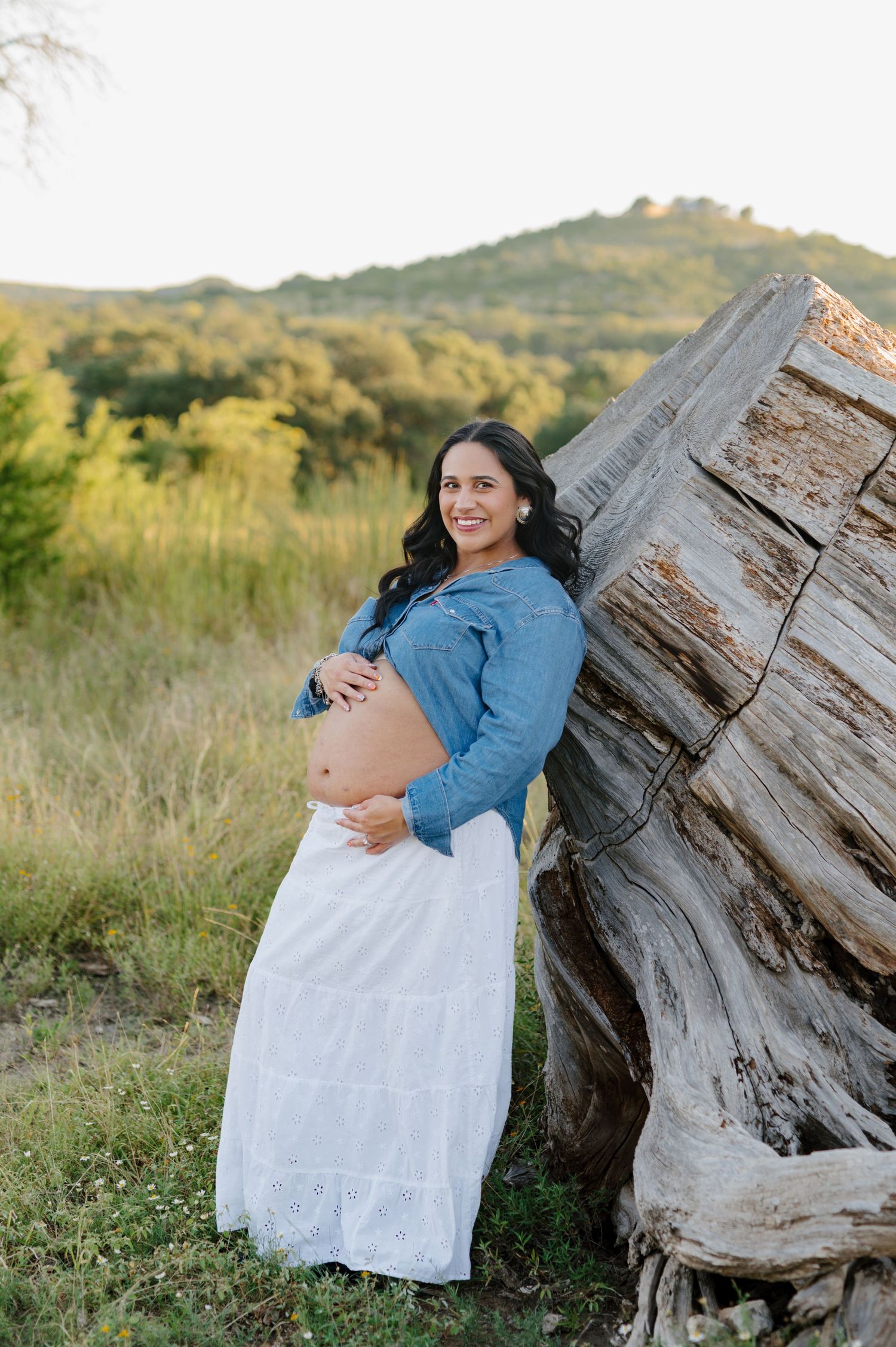 An expectant mother in white dress and denim jacket poses against weathered wood in nature.