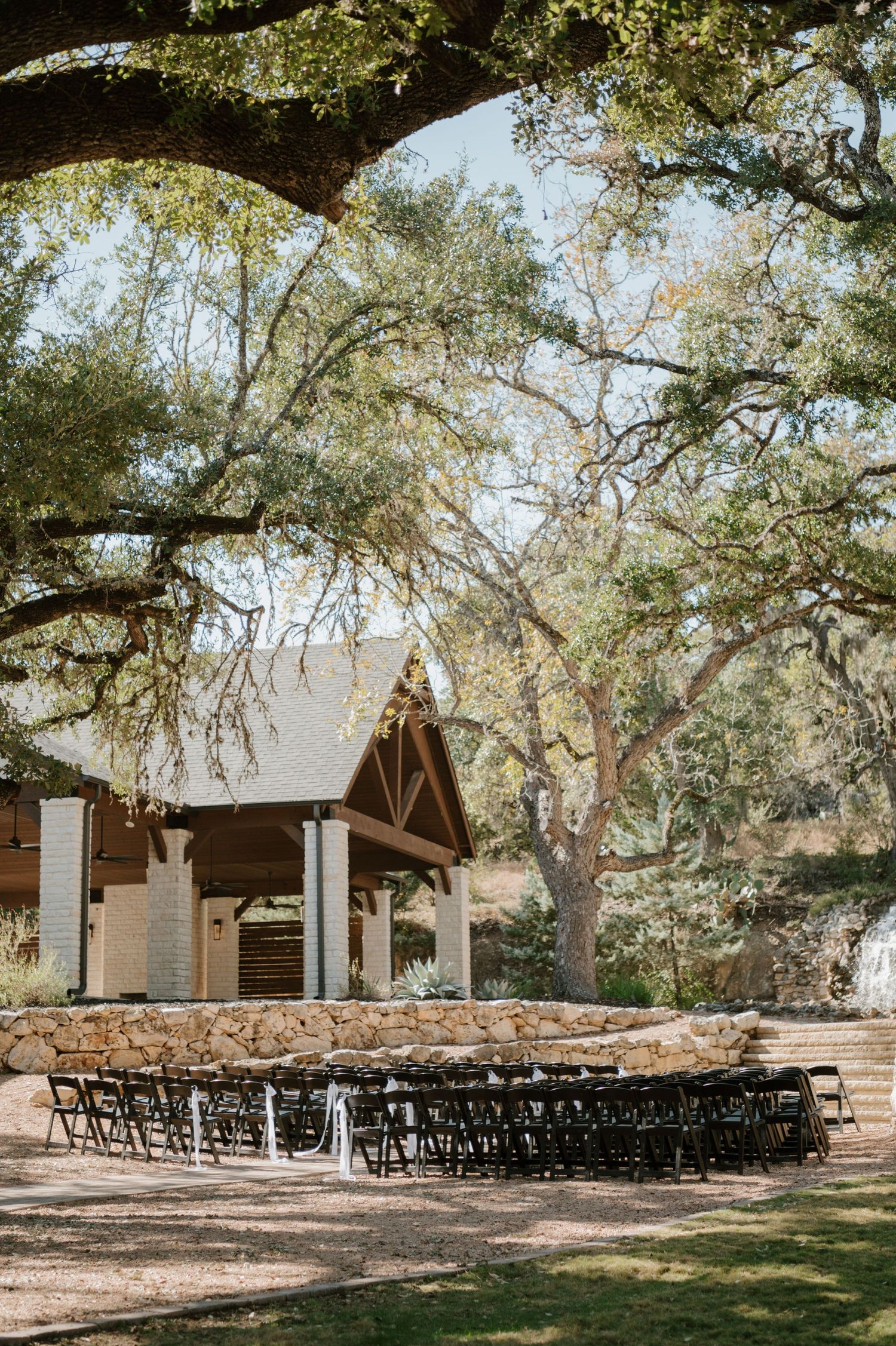 Rustic outdoor wedding ceremony setup with rows of black chairs under large oak trees in the Texas Hill Country.