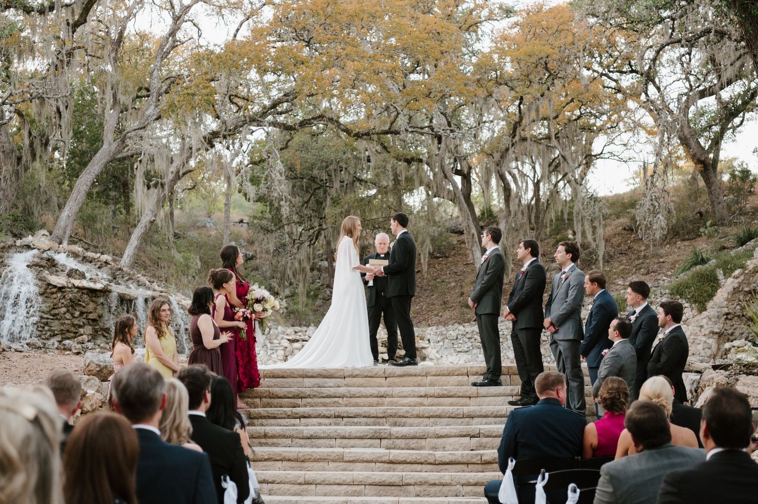 Outdoor wedding ceremony setup with guests gathered under oak trees draped in burgundy floral arrangements.