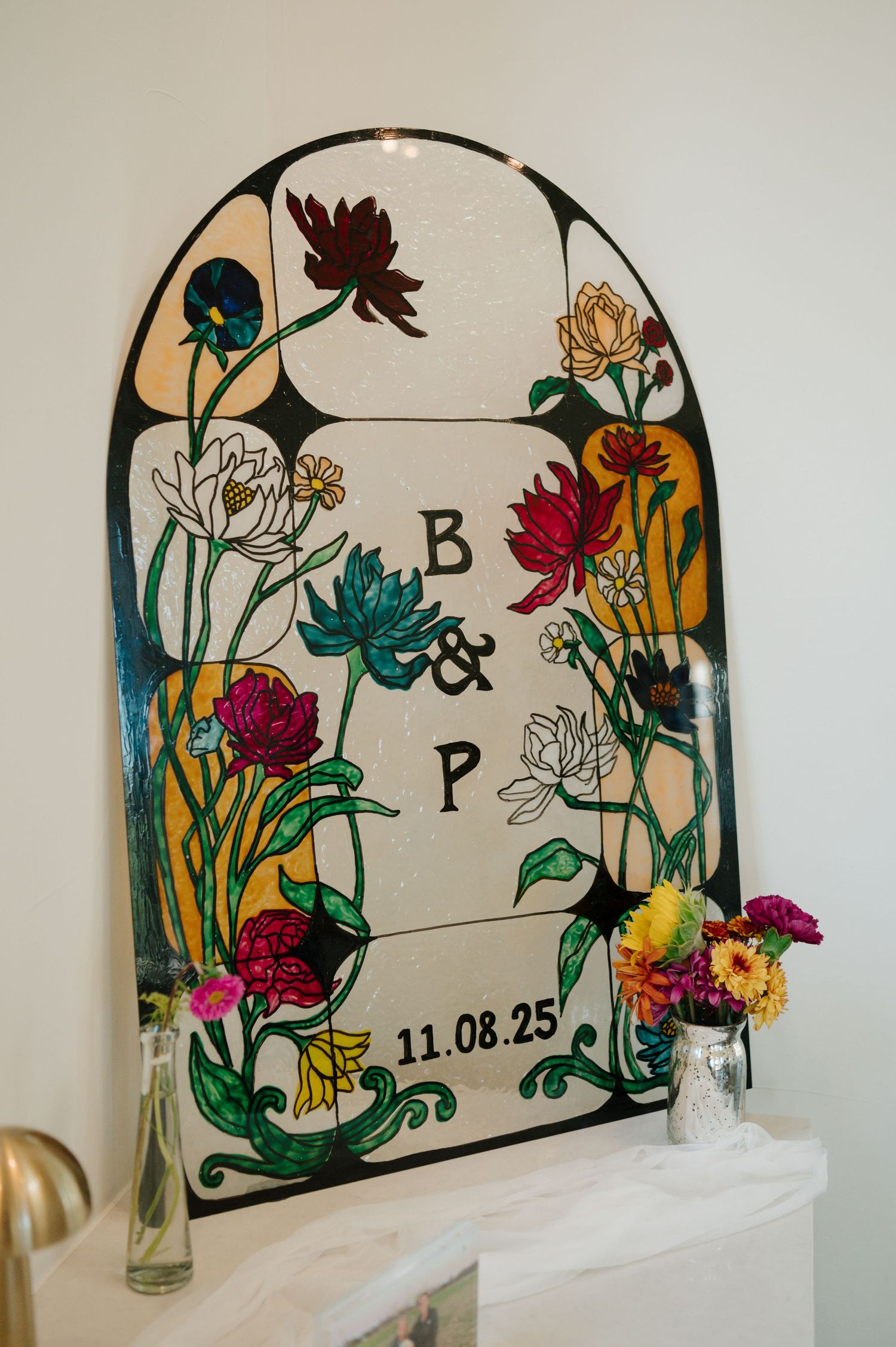 Stained glass art piece featuring colorful floral designs and initials B&P with a date of 11.08.25 in an arched frame.