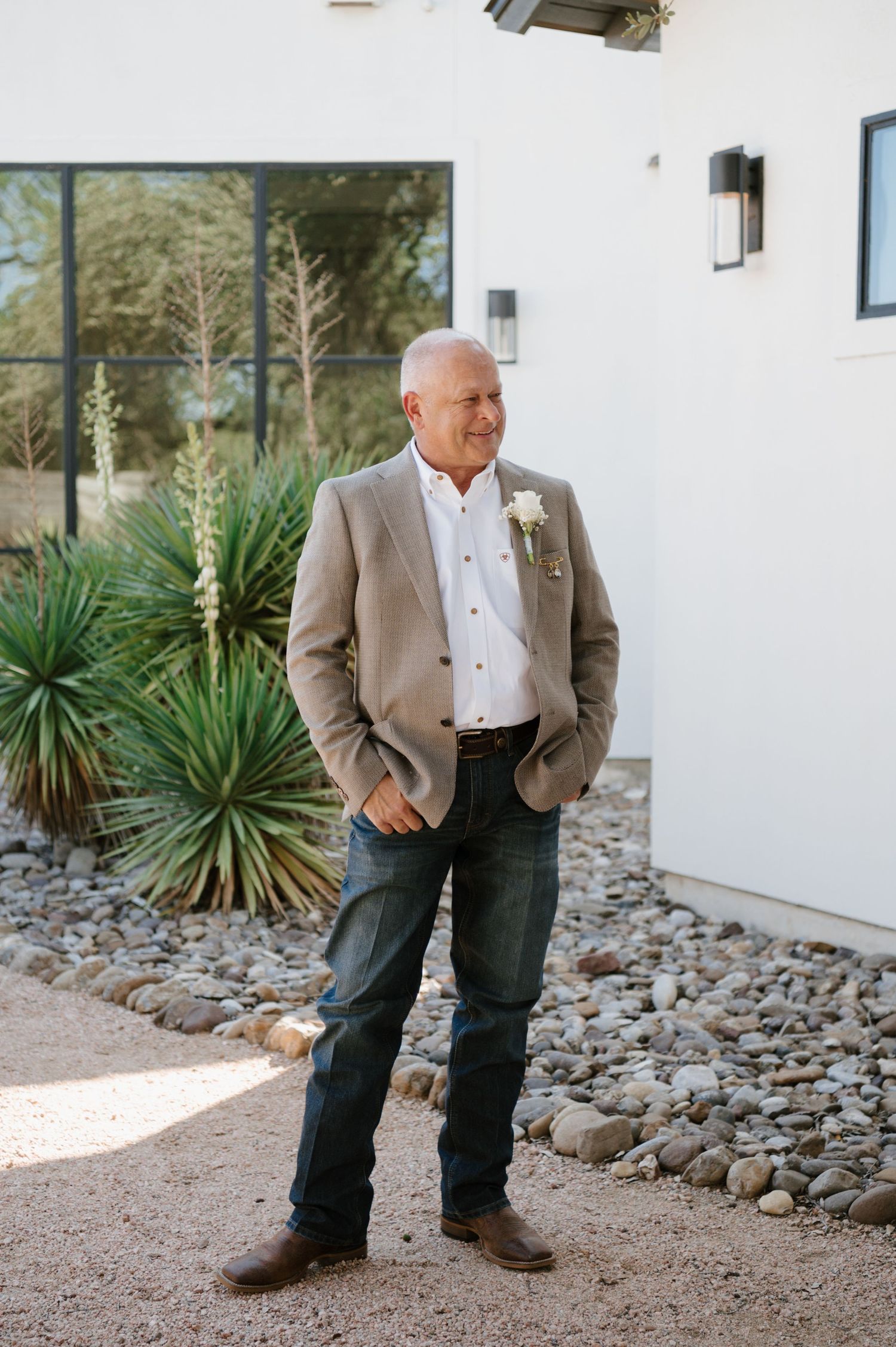 A person wearing a tan blazer and white shirt stands casually in front of a white building with desert landscaping.