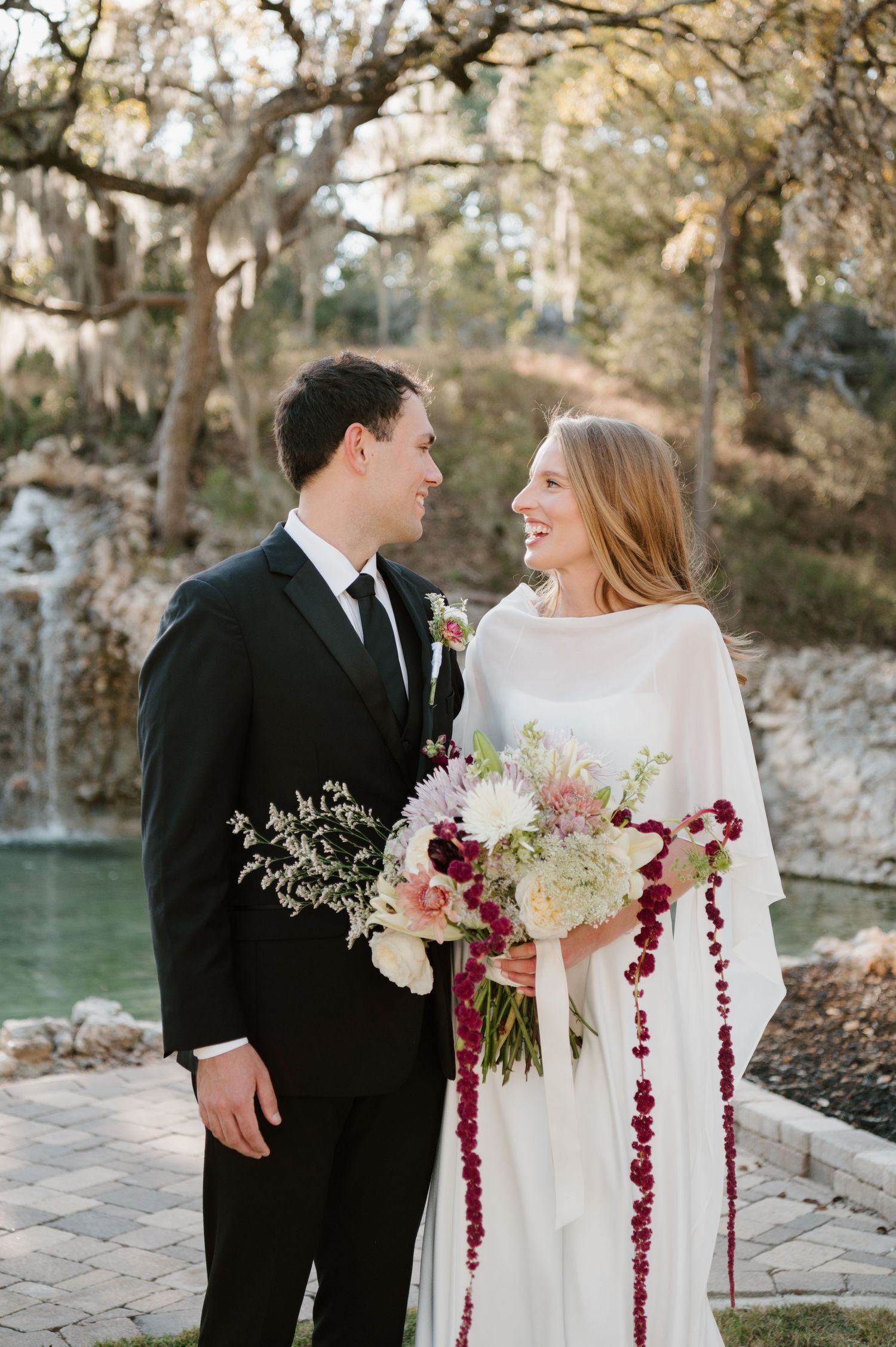 Newlyweds pose with elegant white and burgundy floral bouquet against natural stone backdrop near water.