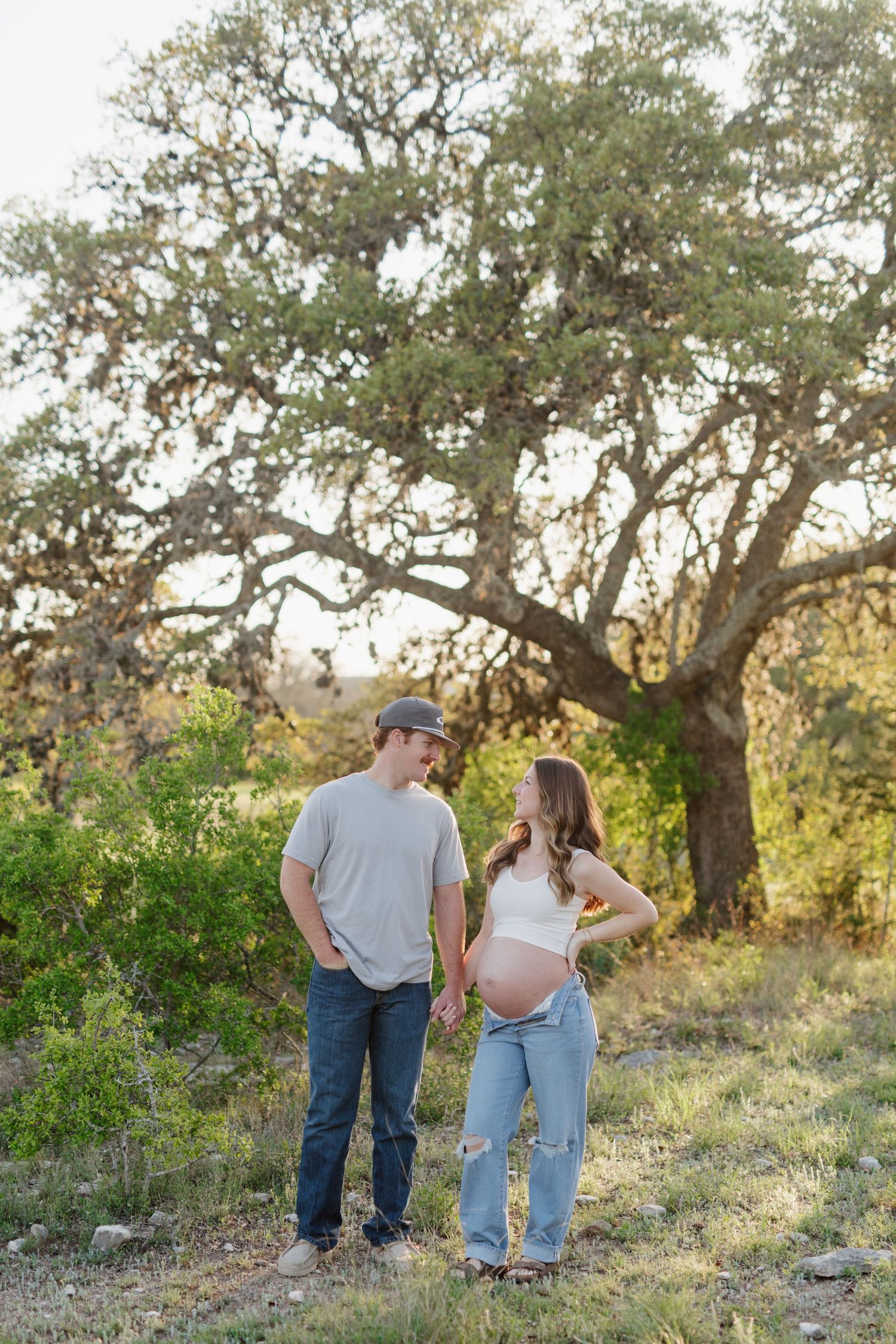 A couple stands together under a large oak tree in a scenic outdoor setting during sunset.
