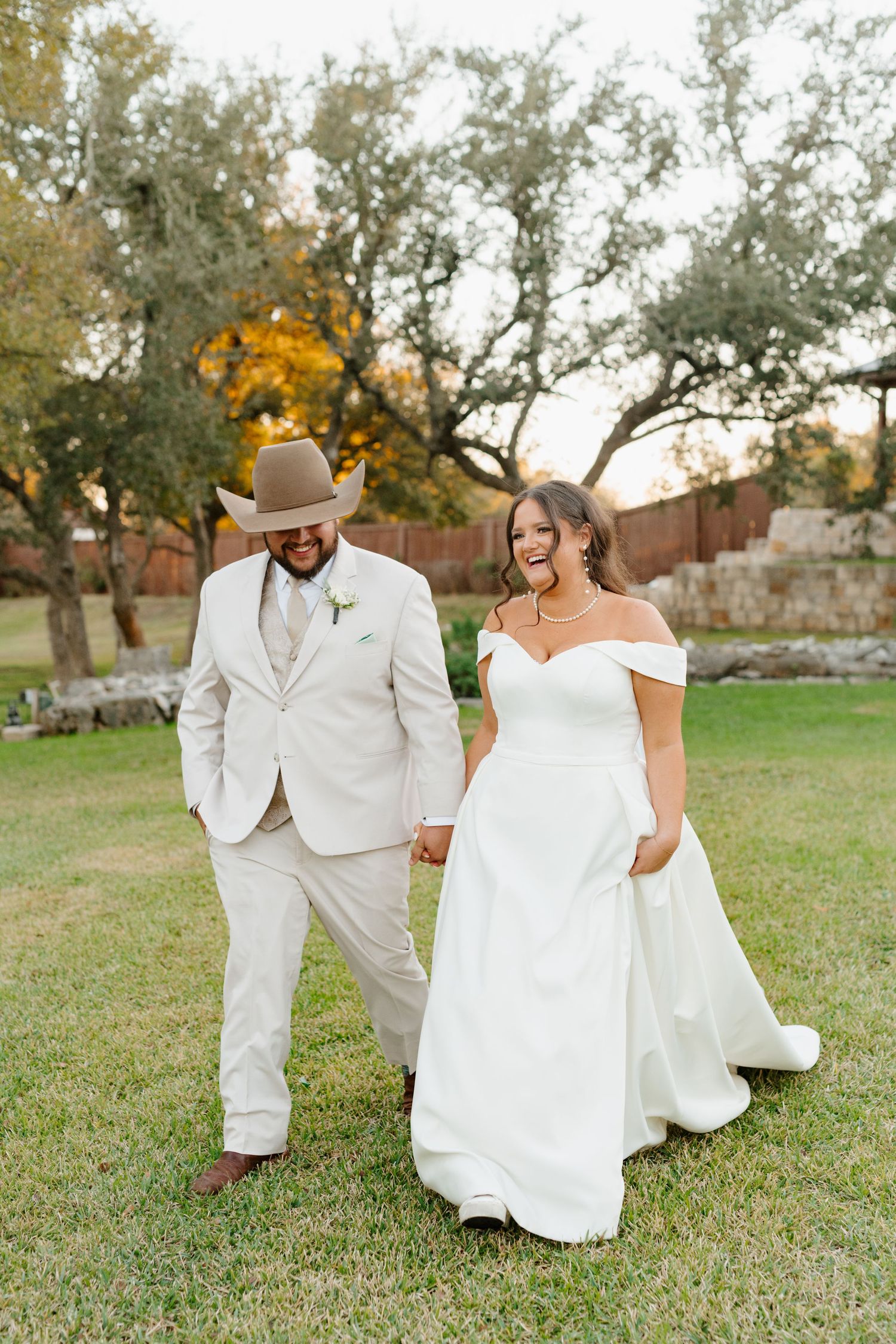 A couple walks hand in hand outdoors on their wedding day, wearing a light beige suit and off-shoulder white gown.
