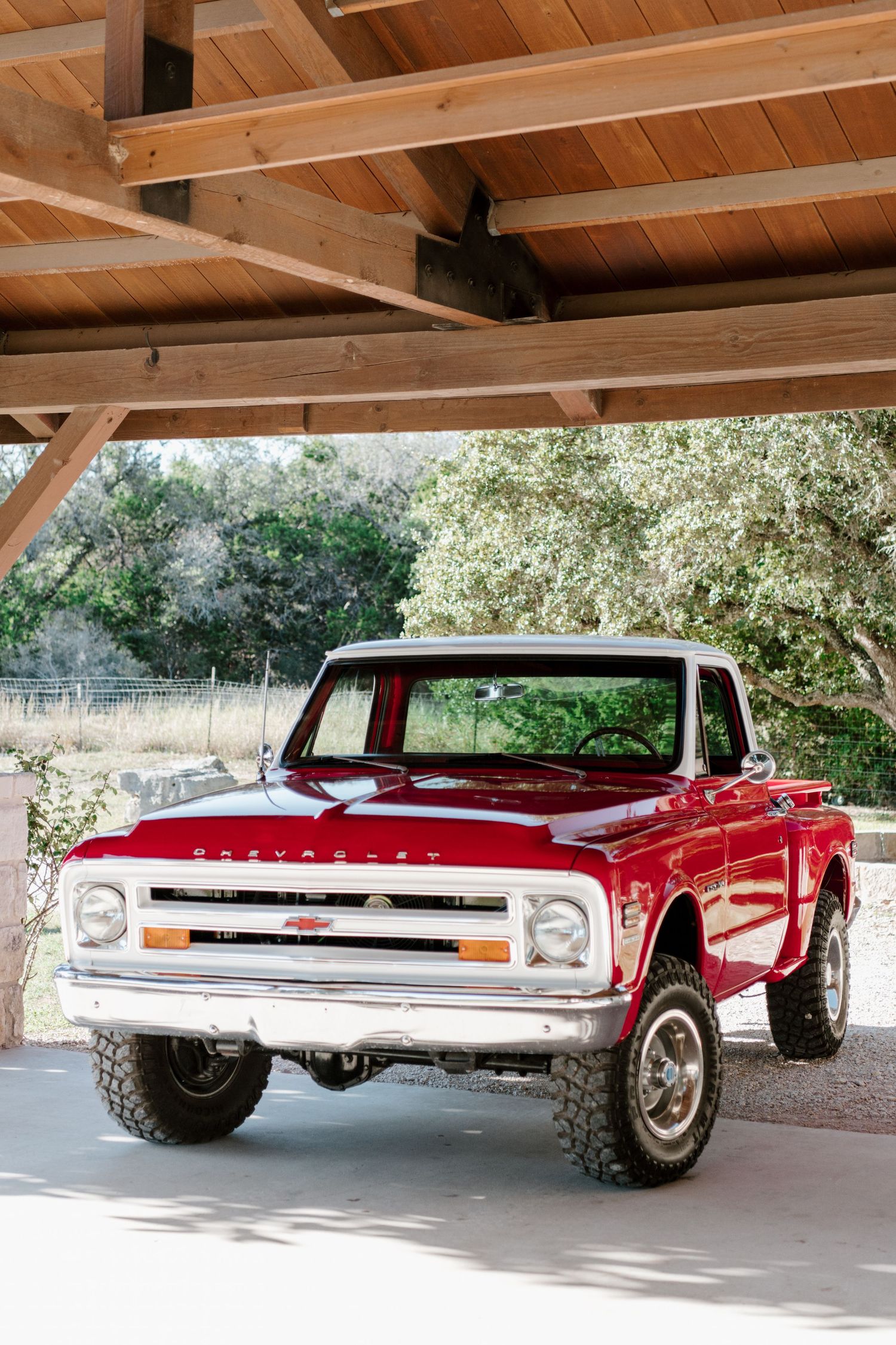 A classic red Chevrolet C10 pickup truck with lifted suspension parked under a wooden pergola structure.